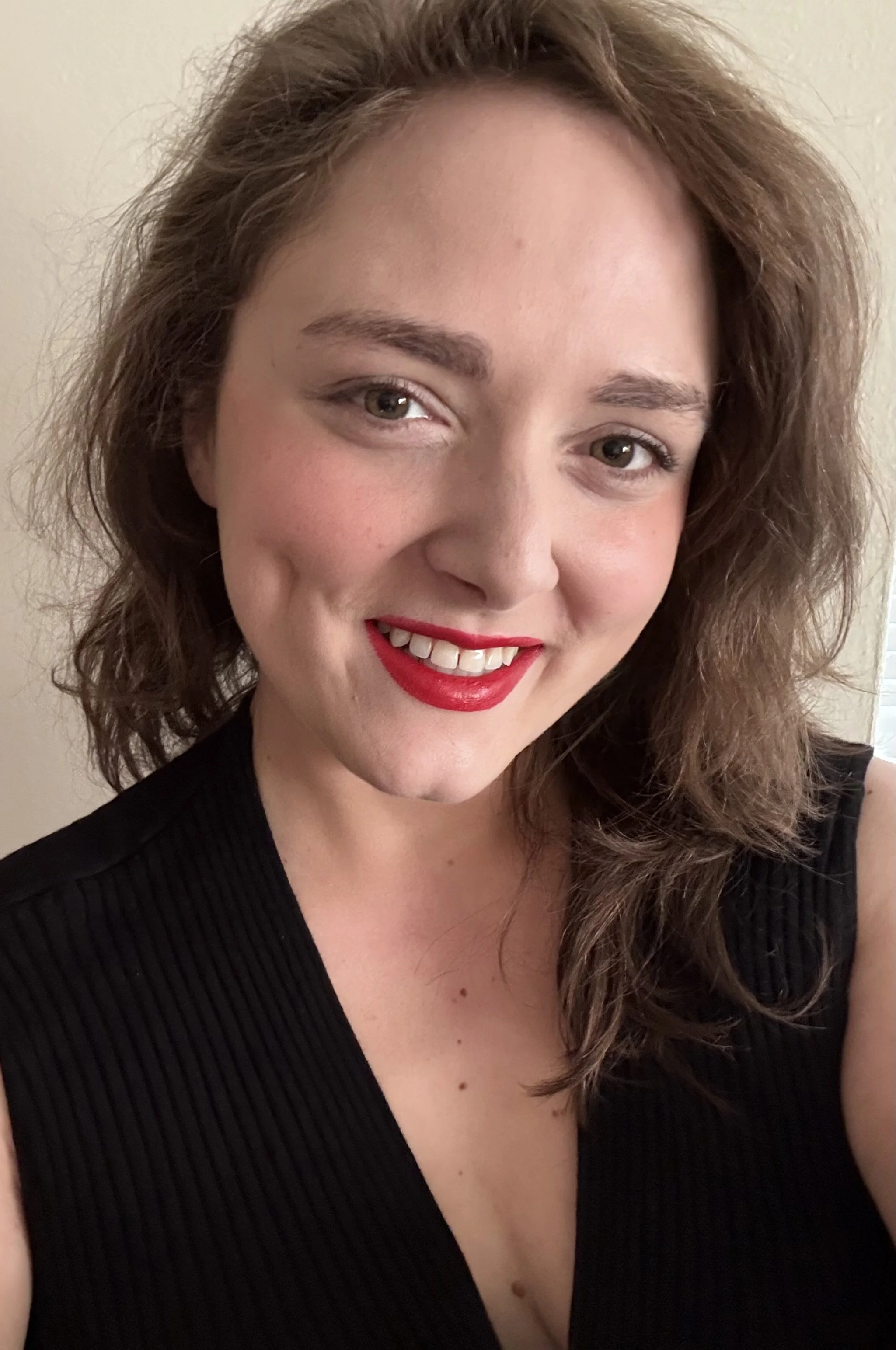 A woman with shoulder-length wavy brown hair, wearing red lipstick and a black sleeveless top, smiling at the camera.