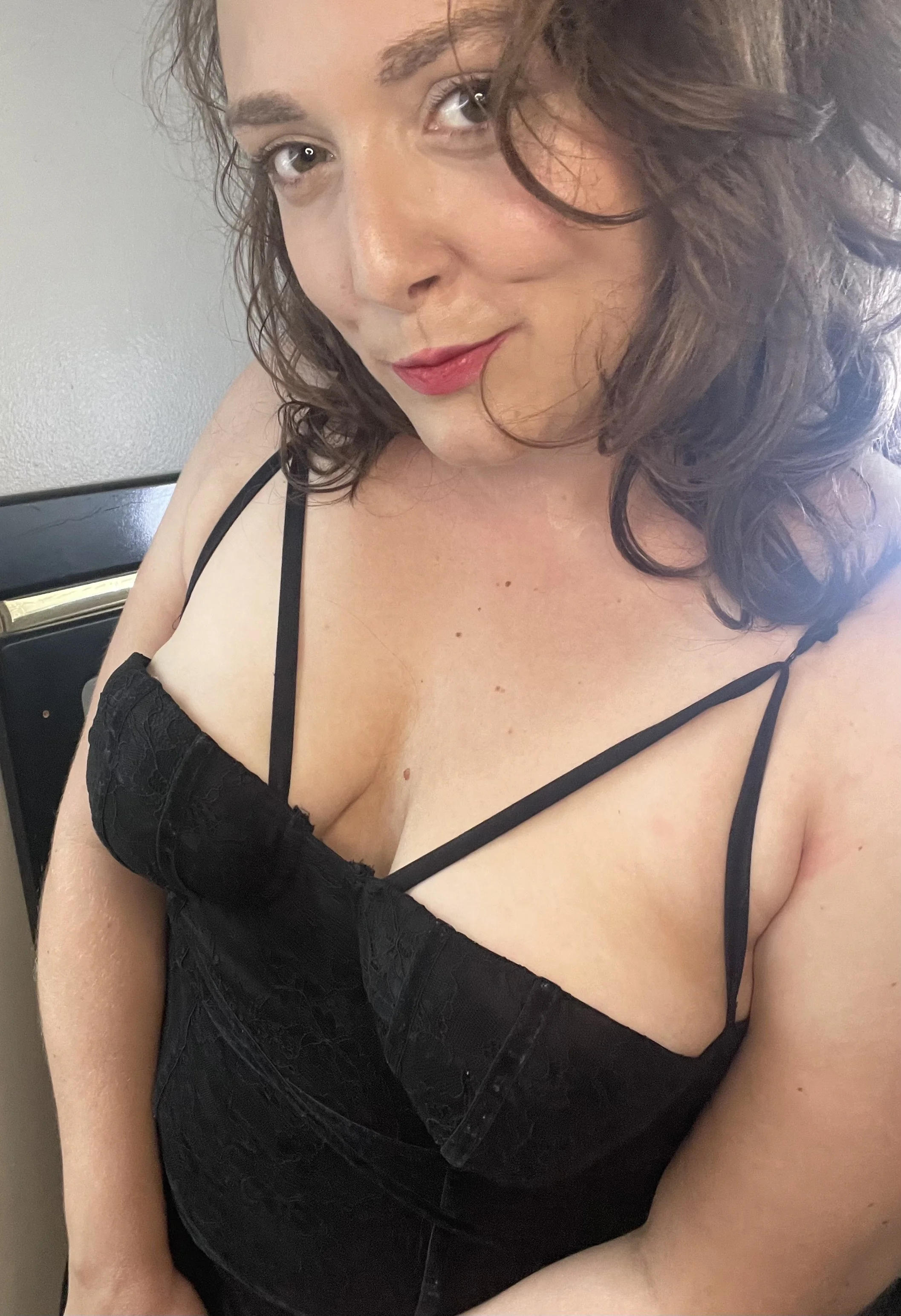 A woman with curly brown hair wearing a black dress with spaghetti straps and lace detailing. She is smiling softly and looking at the camera.