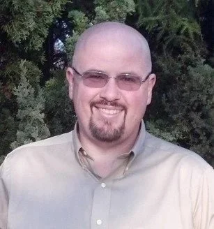 A man with glasses and a goatee, smiling outdoors in front of trees, wearing a beige shirt.