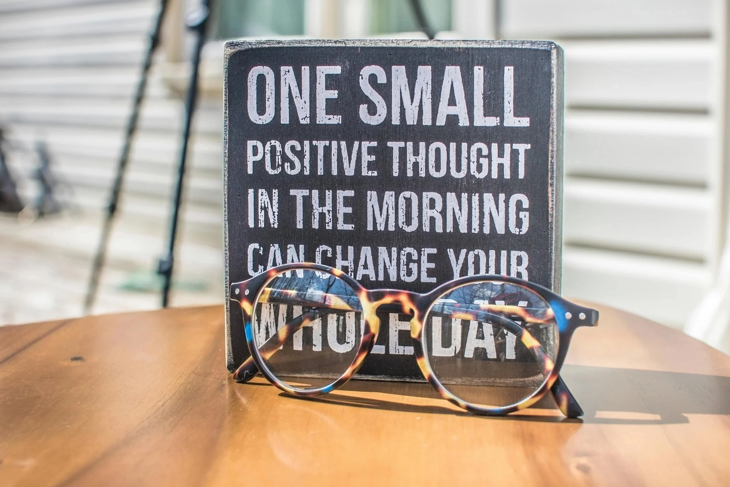 A small blackboard sign with motivational quote standing on a wooden table, accompanied by round tortoiseshell glasses.