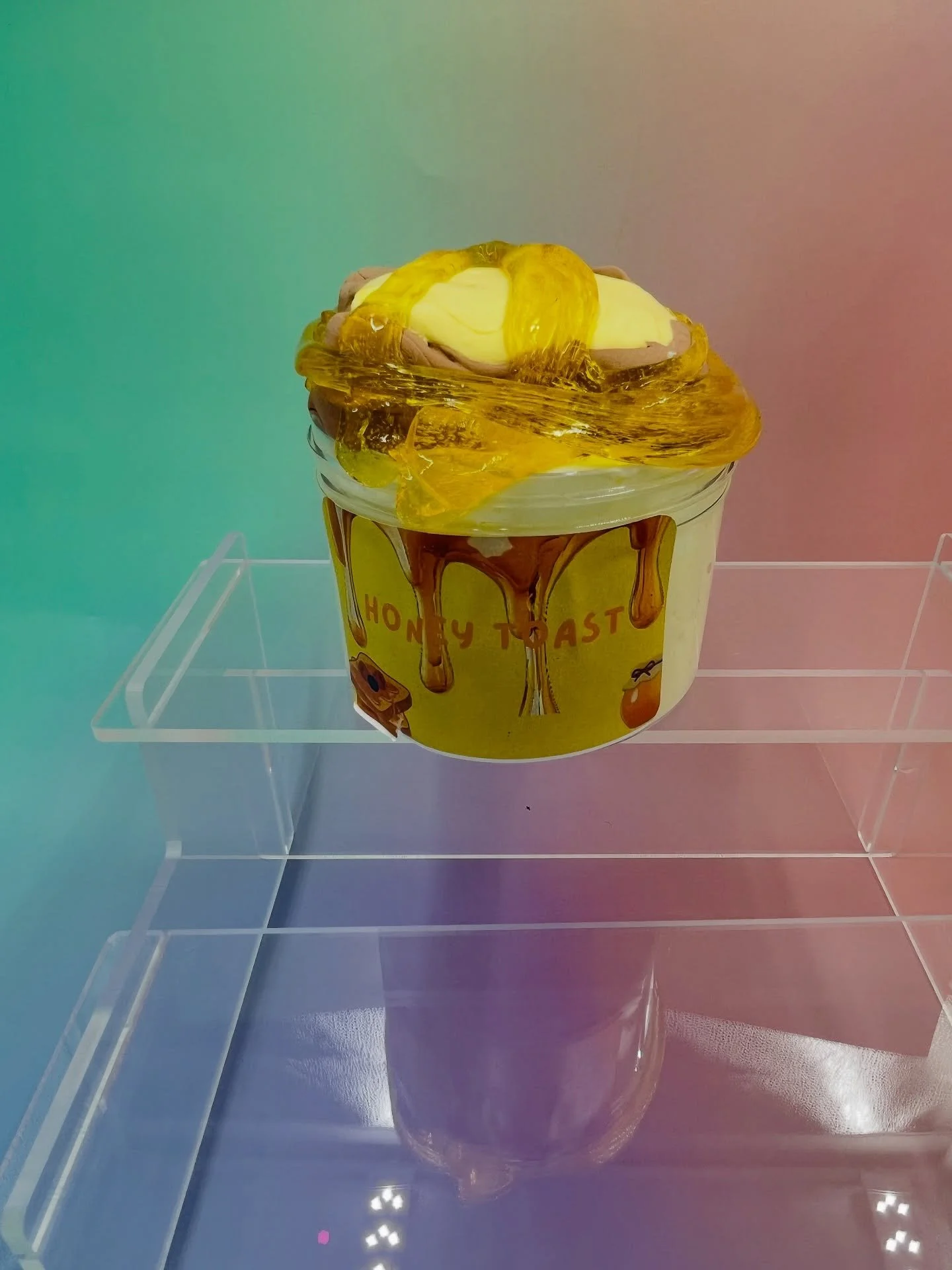 A honey toast dessert with a yellow bowl labeled ’HONEY TOAST’, topped with a scoop of ice cream, honey, and chocolate syrup, displayed on a clear stand.