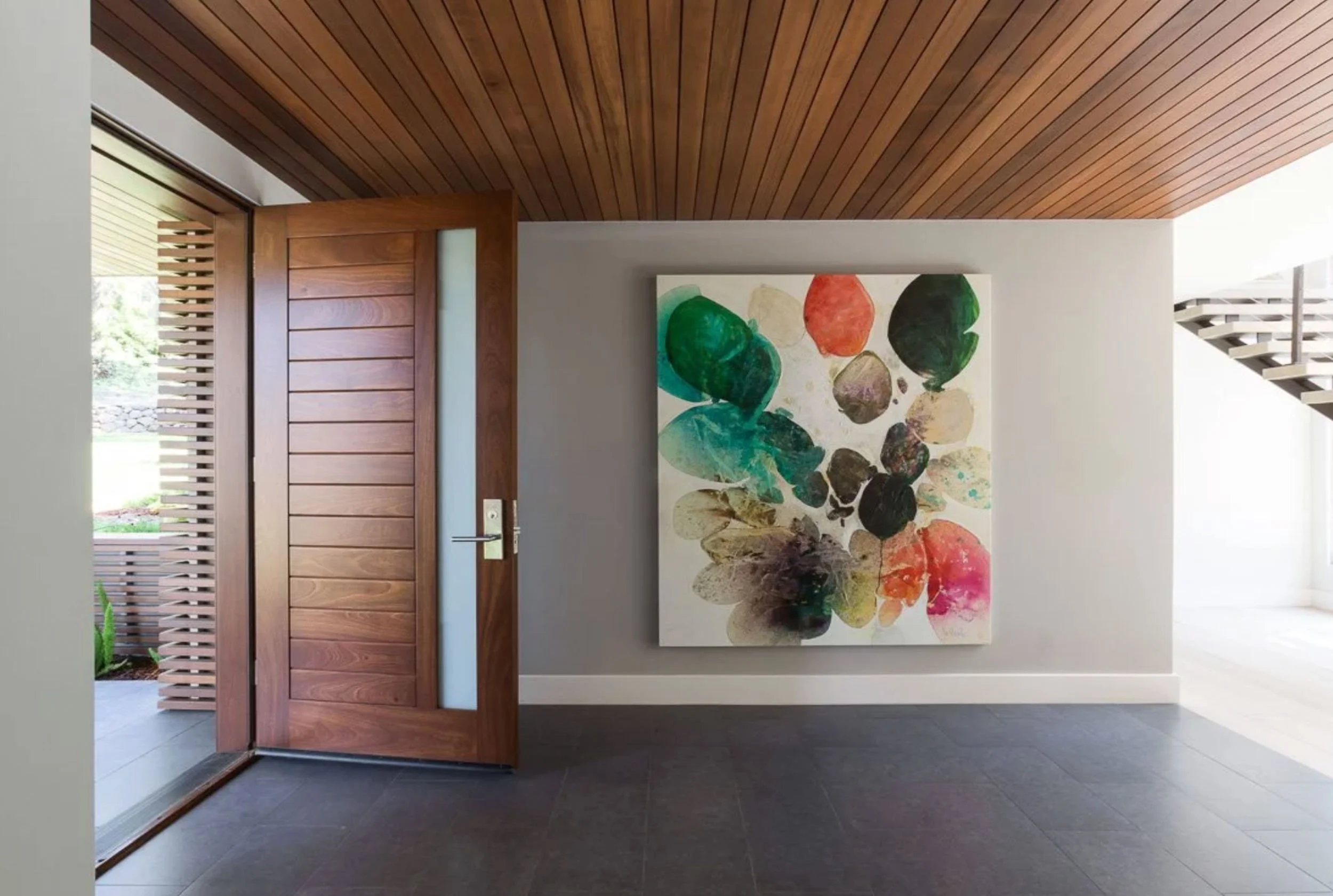 A colorful abstract painting of translucent organic shapes in green, orange, teal, and magenta hangs in an entryway, set off by a wood door and slatted wood ceiling.