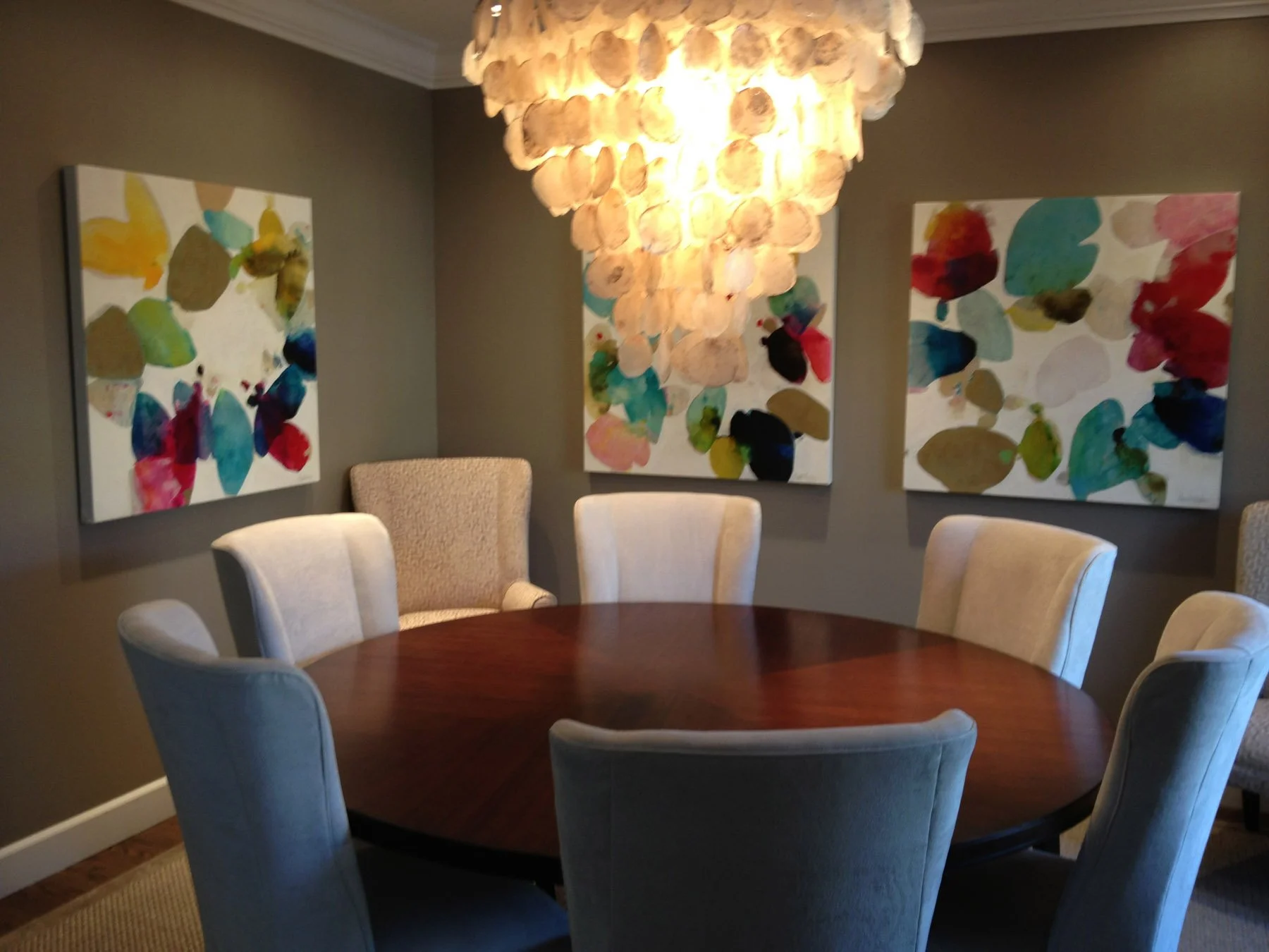 Three colorful abstract paintings of organic, petal-like shapes in teal, magenta, gold, and red line the gray walls of a dining room, above a round wooden table ringed with upholstered chairs.