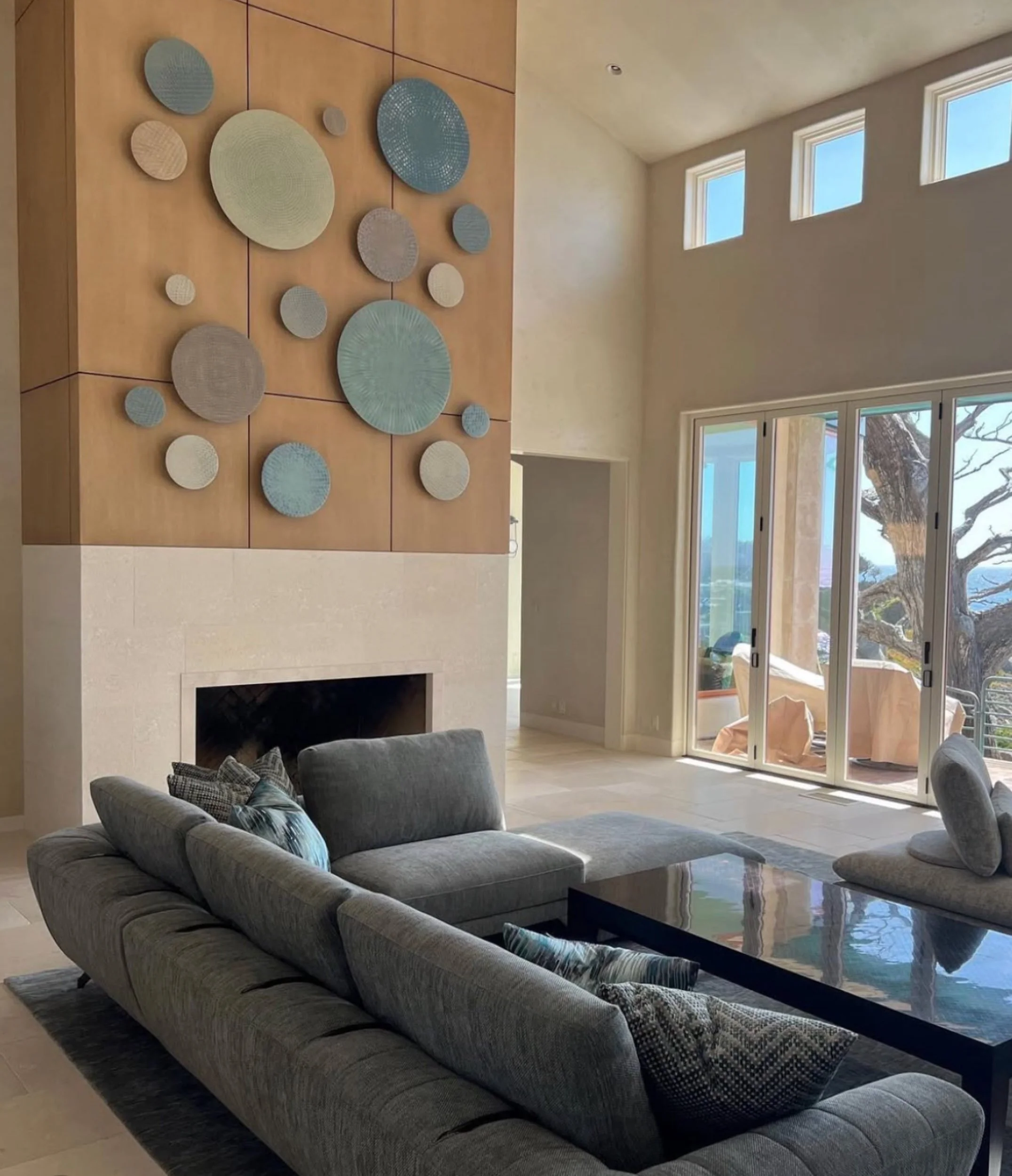 A cluster of round ceramic wall discs in muted blue, green, and gray, mounted on a wood-panel wall above the fireplace of a living room with tall windows overlooking the trees outside.