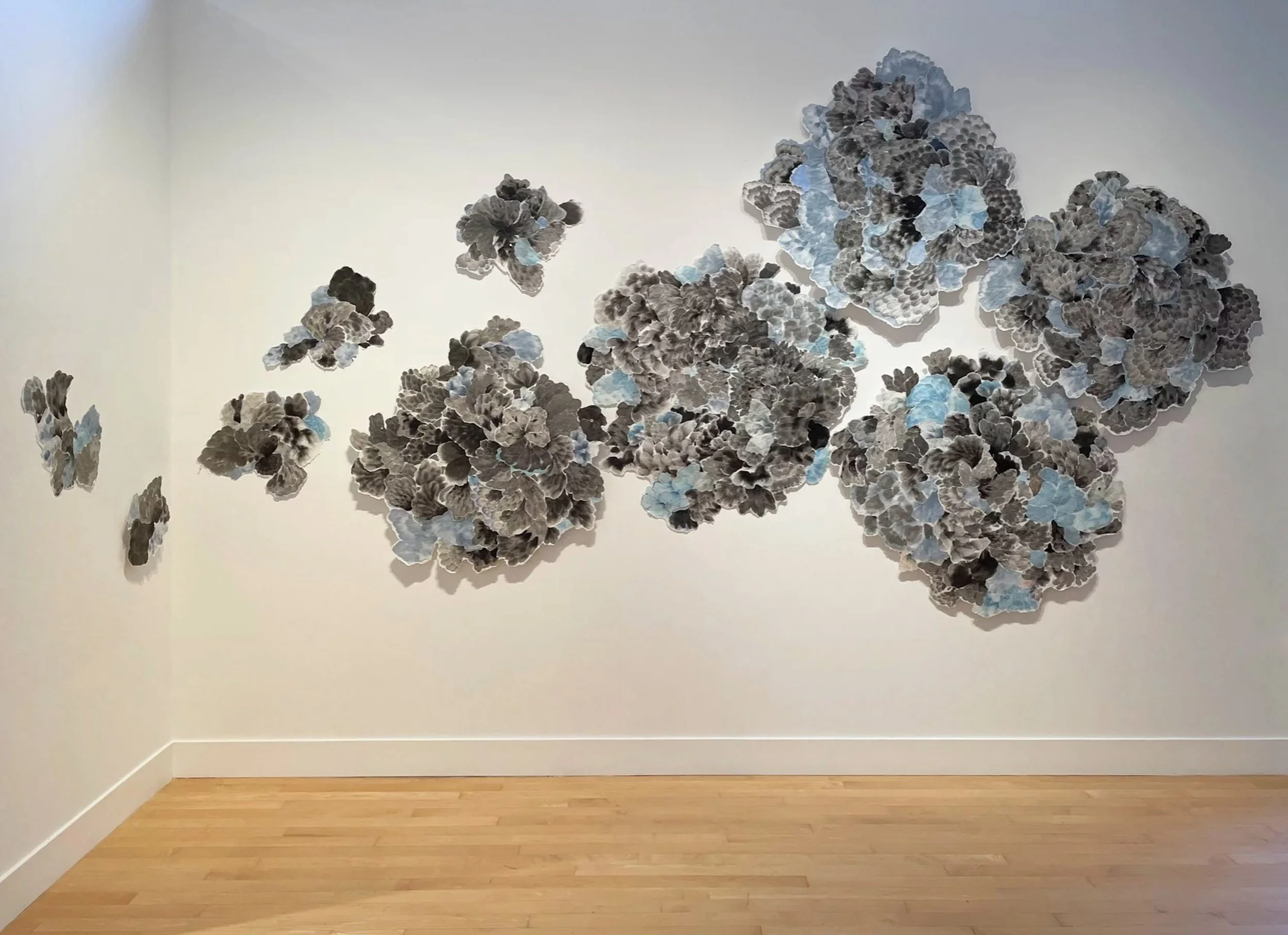 A large abstract wall sculpture of layered paper, resembling clusters of blossoms in black, gray, and pale blue, spread across a white gallery wall.