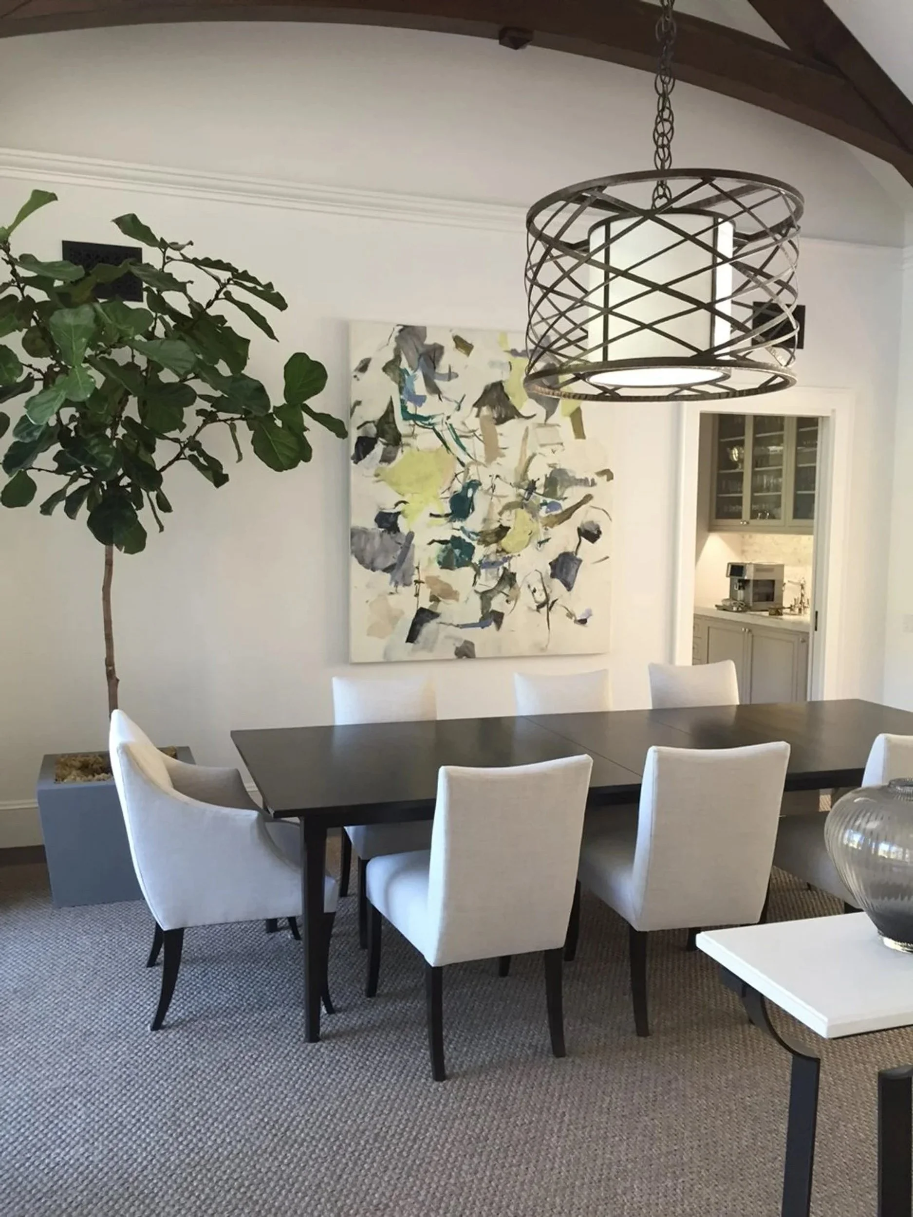 A colorful gestural abstract painting in yellow, blue, and green hangs on the wall of a dining room, behind a dark wood table ringed with white upholstered chairs.