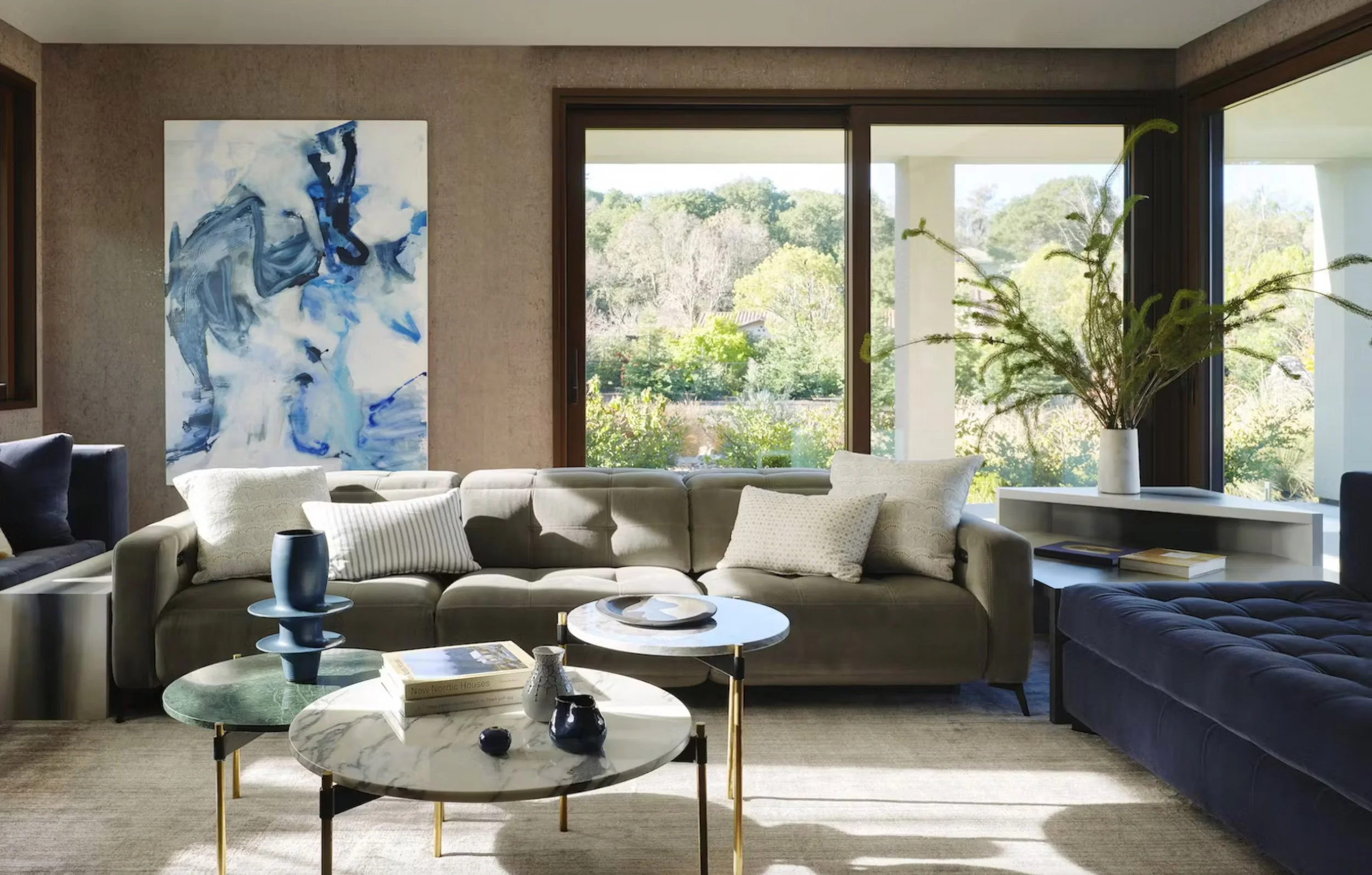 A large vertical abstract painting in fluid blue and white hangs above the sofa in a sunlit living room, with round marble tables and windows opening onto green trees.