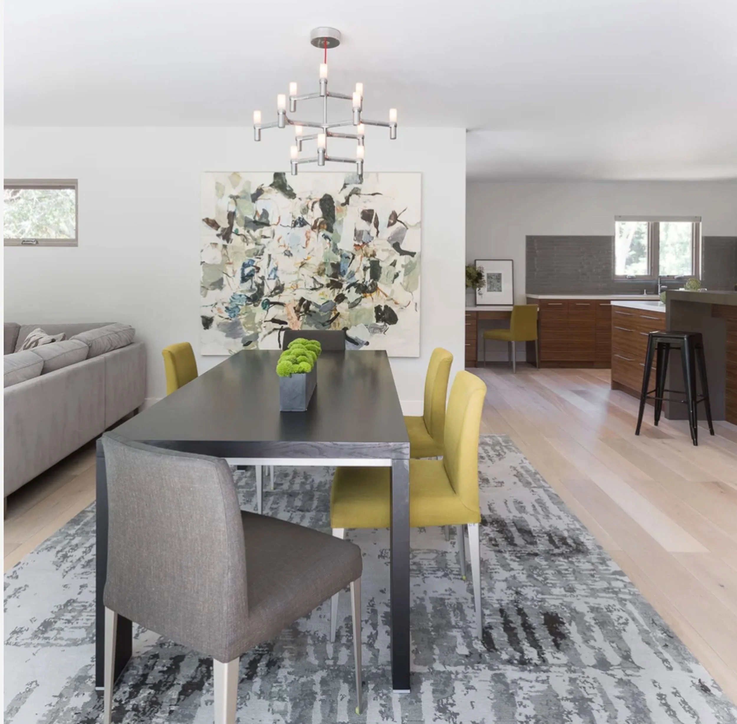 A large abstract painting with gestural, foliage-like marks in muted gray, blue, green, and gold anchors the white wall of a modern dining room, above a gray table and yellow upholstered chairs.
