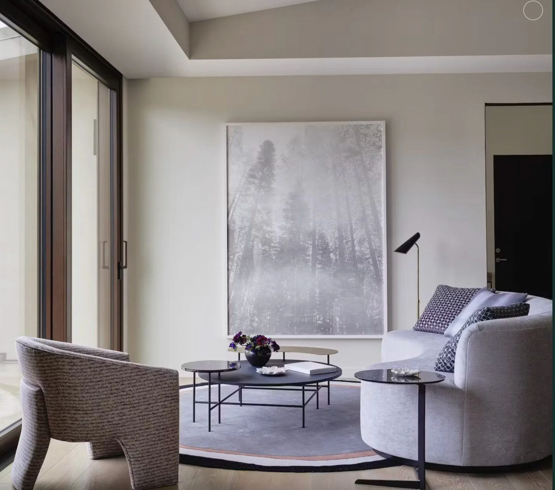 A large black-and-white misty forest artwork anchors a modern living room, set above round coffee tables between a curved woven armchair and a grey sofa with floor-to-ceiling glass doors beyond.