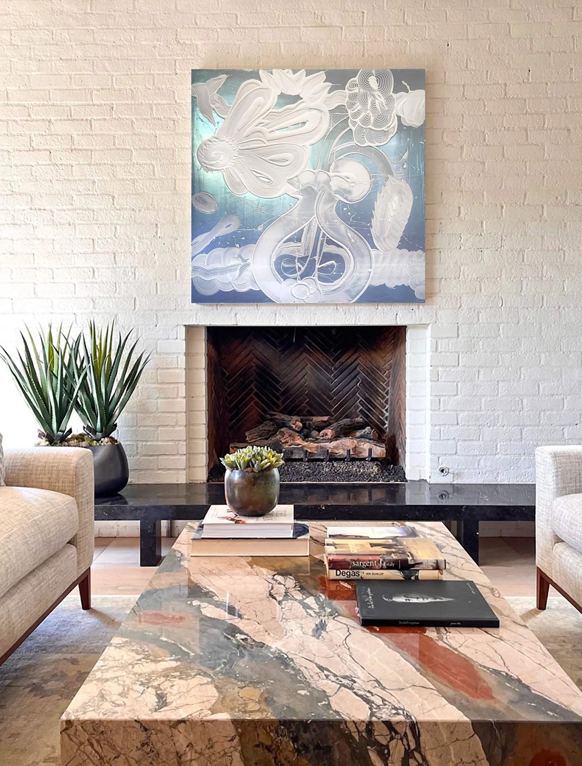 A large abstract painting in blue and gray with swirling white line-work hangs above the fireplace on a white brick wall, with armchairs and a boldly veined marble coffee table below.