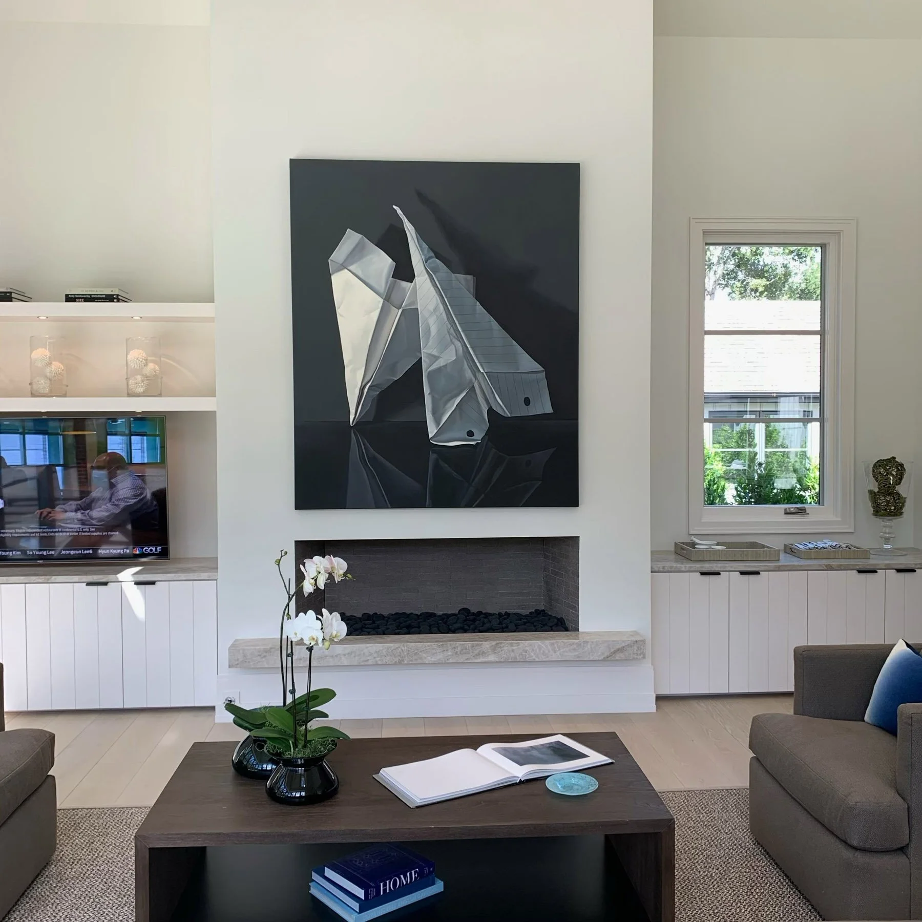 A photorealist painting of folded paper airplanes, set on a black ground hangs above the white fireplace of a modern living room, with orchids on the dark wood coffee table below.