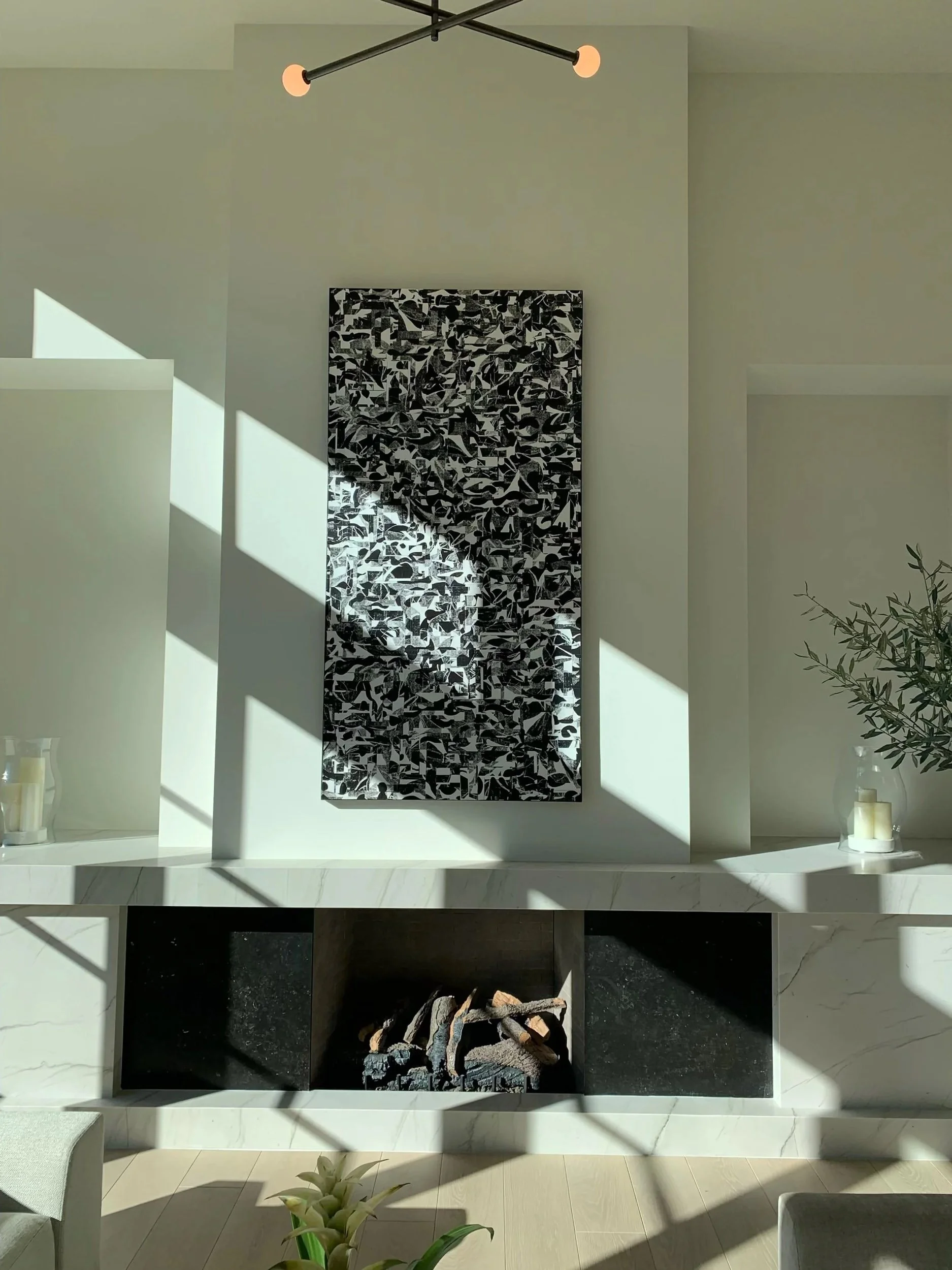 A modern fireplace with black and white abstract artwork above it, candles on side shelves, and sunlight casting shadows in a clean, minimalist living space.