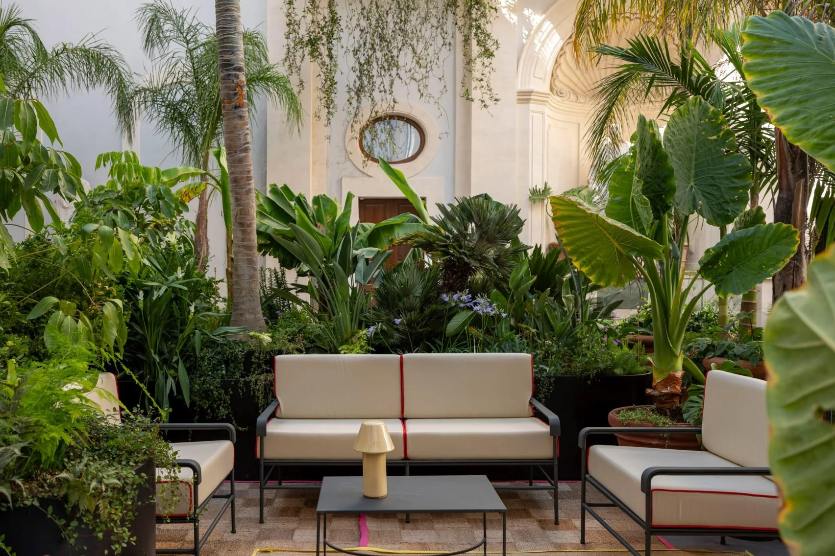 Indoor seating area with modern white sofas, a black coffee table with a beige vase, and lush tropical plants in pots, set against a background of a white wall with architectural details and a window.