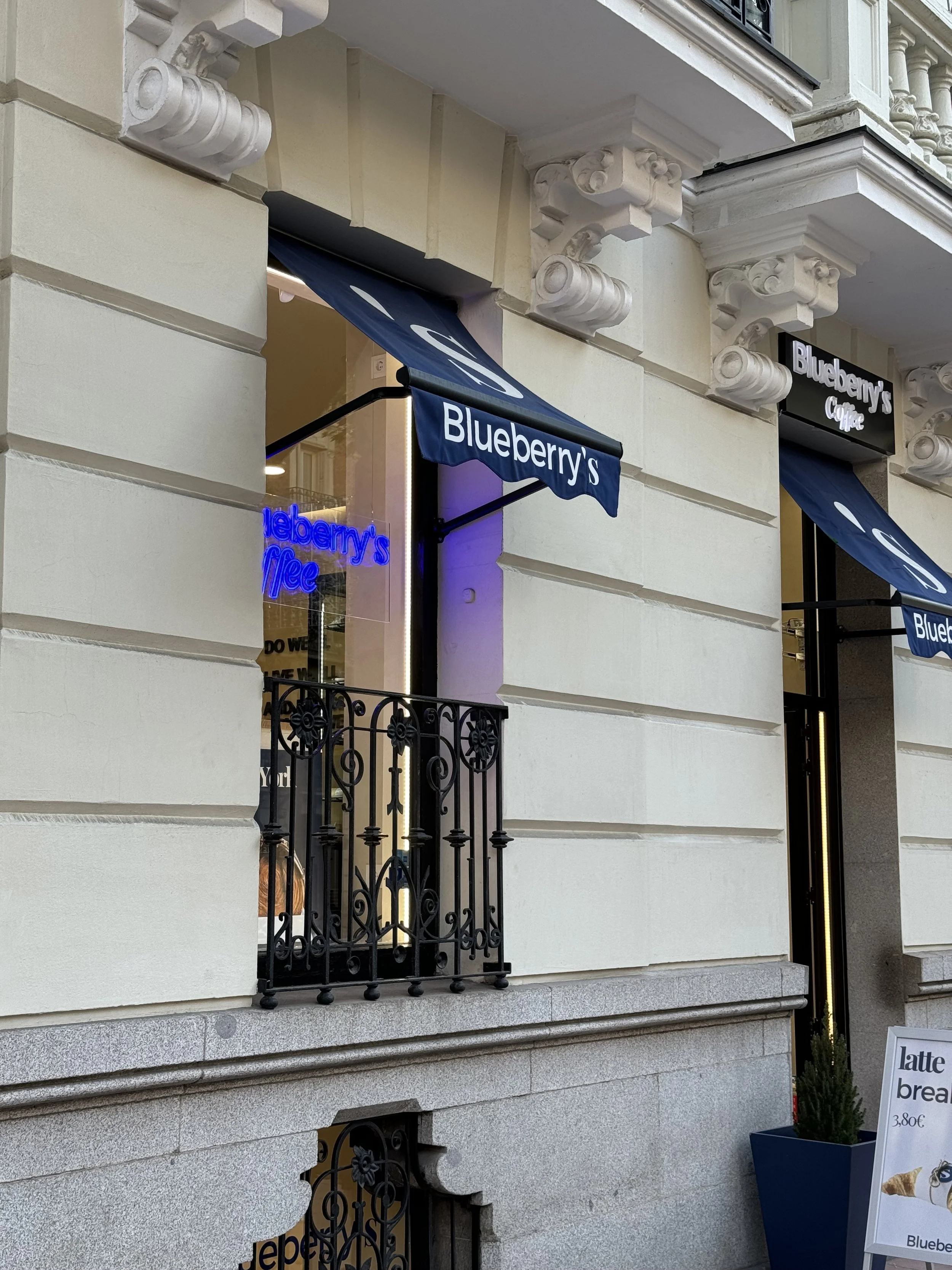 Exterior of a cafe named Blueberry's with beige building facade, black ornate balcony, and blue awnings displaying the name. Neon sign inside reads Blueberry's Coffee.