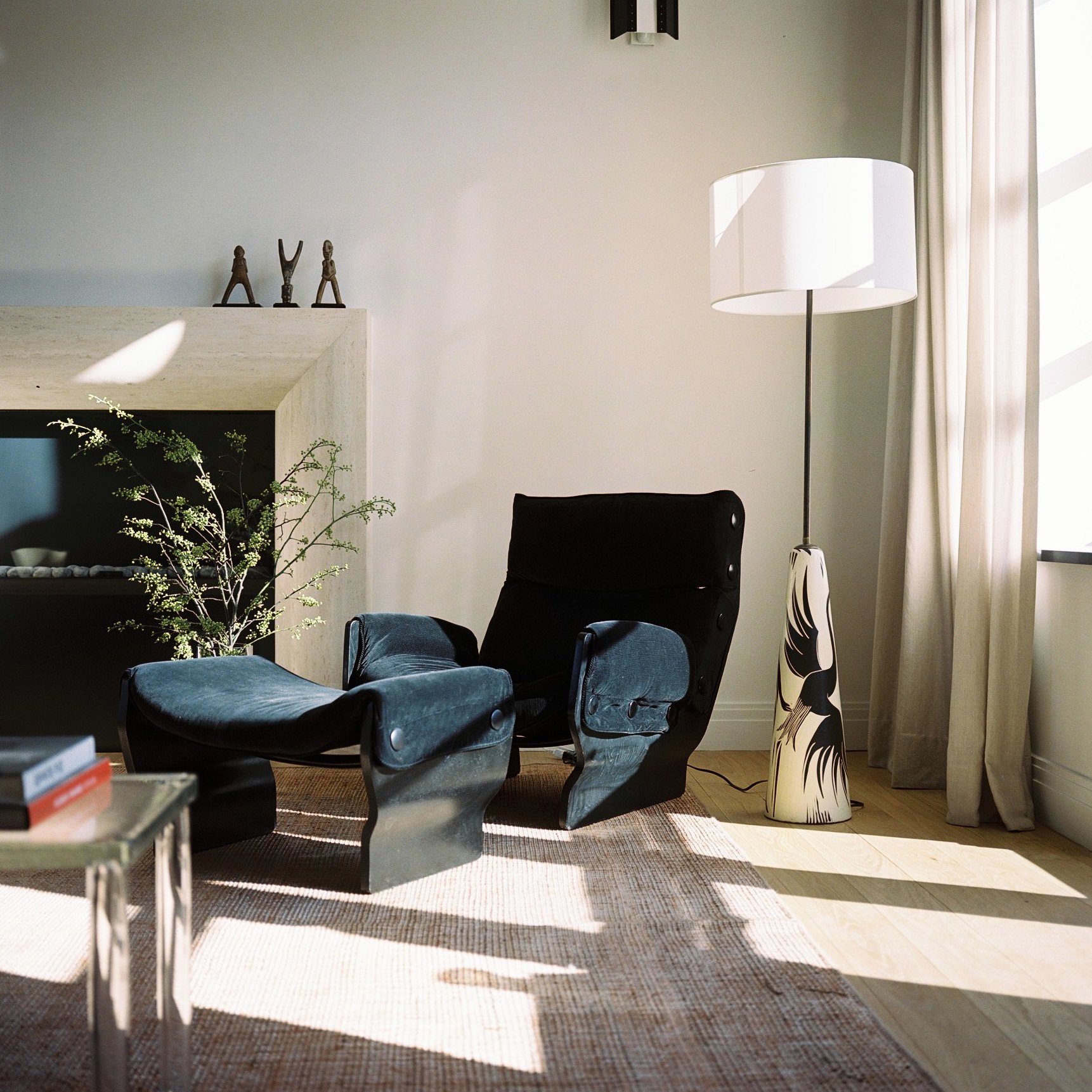 Modern living room with black armchair, tall floor lamp with patterned base, beige curtains, and sunlight through windows.