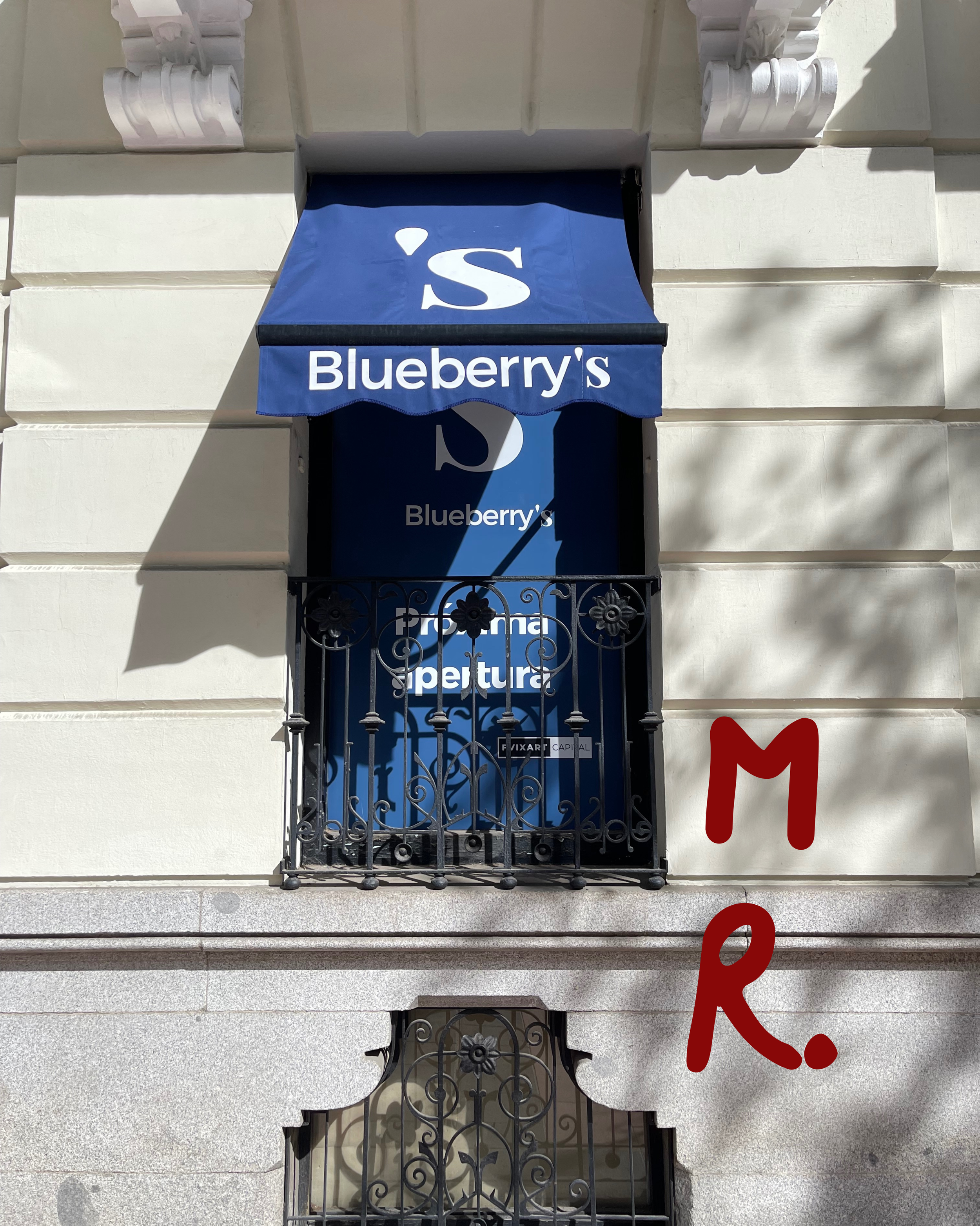 Facade of a building with a blue awning displaying a white S and the word 'Blueberry's'. Below, there is a window with an ornate black iron railing and a blue sheet or sign behind it. To the right, on the building's wall, is the red text 'M R.'