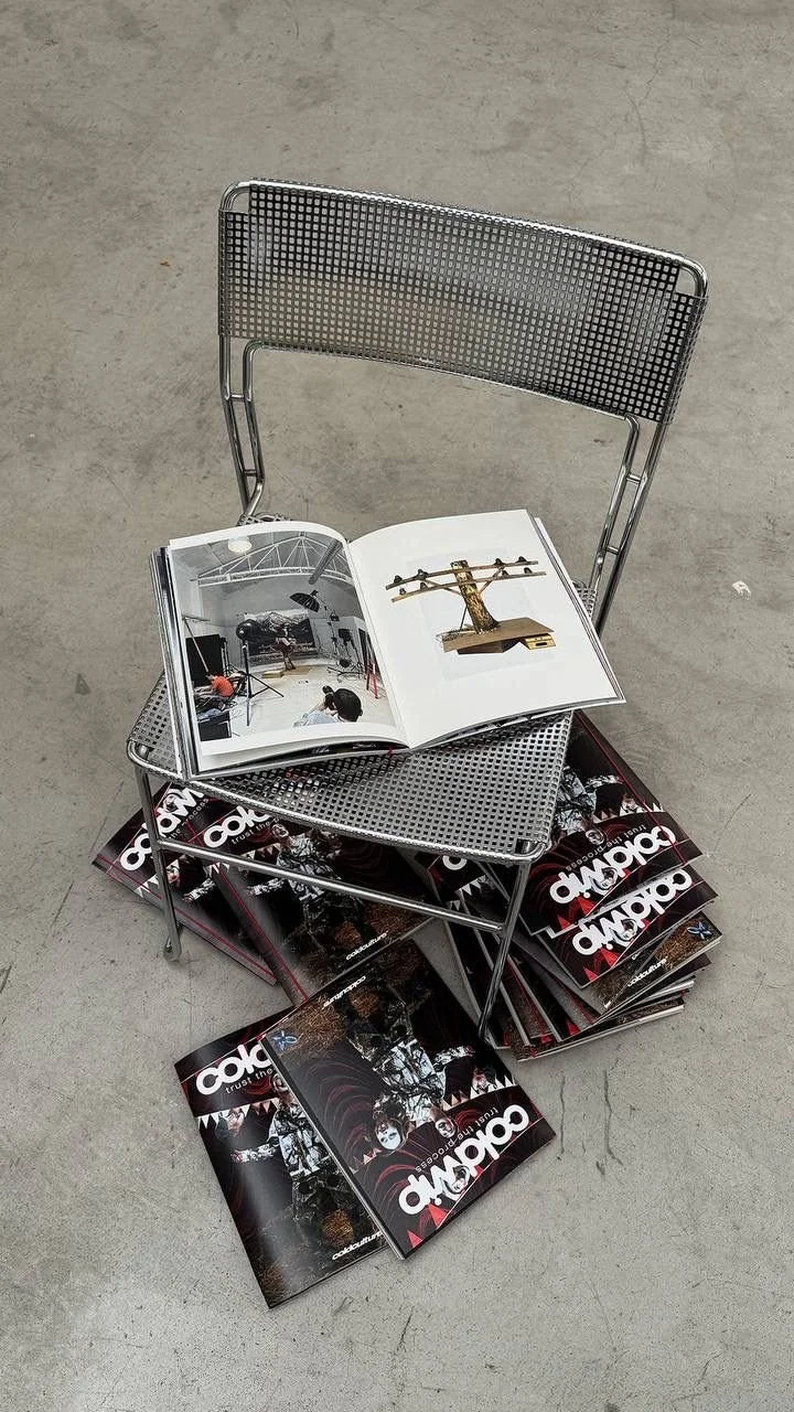 A metal chair with magazines underneath, an open magazine on the seat displaying pictures of an art installation, and another open magazine on top showing a miniature sculpture. The floor is gray concrete.