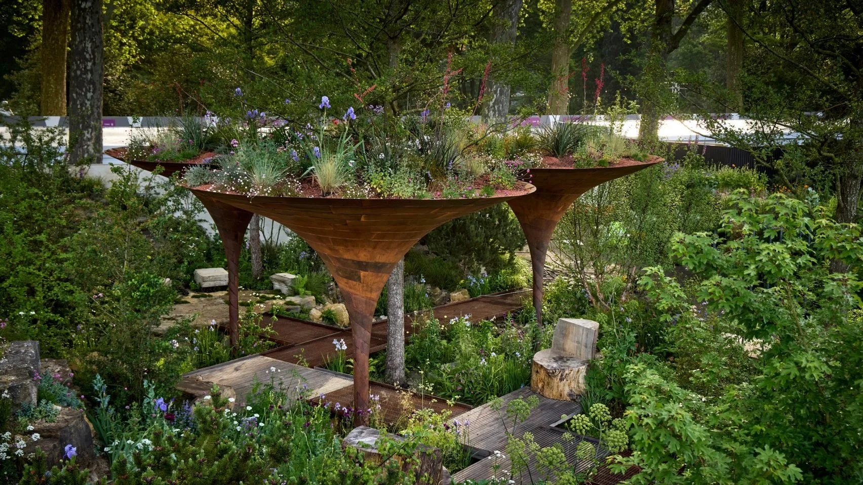 A garden featuring elevated wooden planter beds shaped like upside-down cones, filled with various plants and flowers, surrounded by lush greenery and trees.