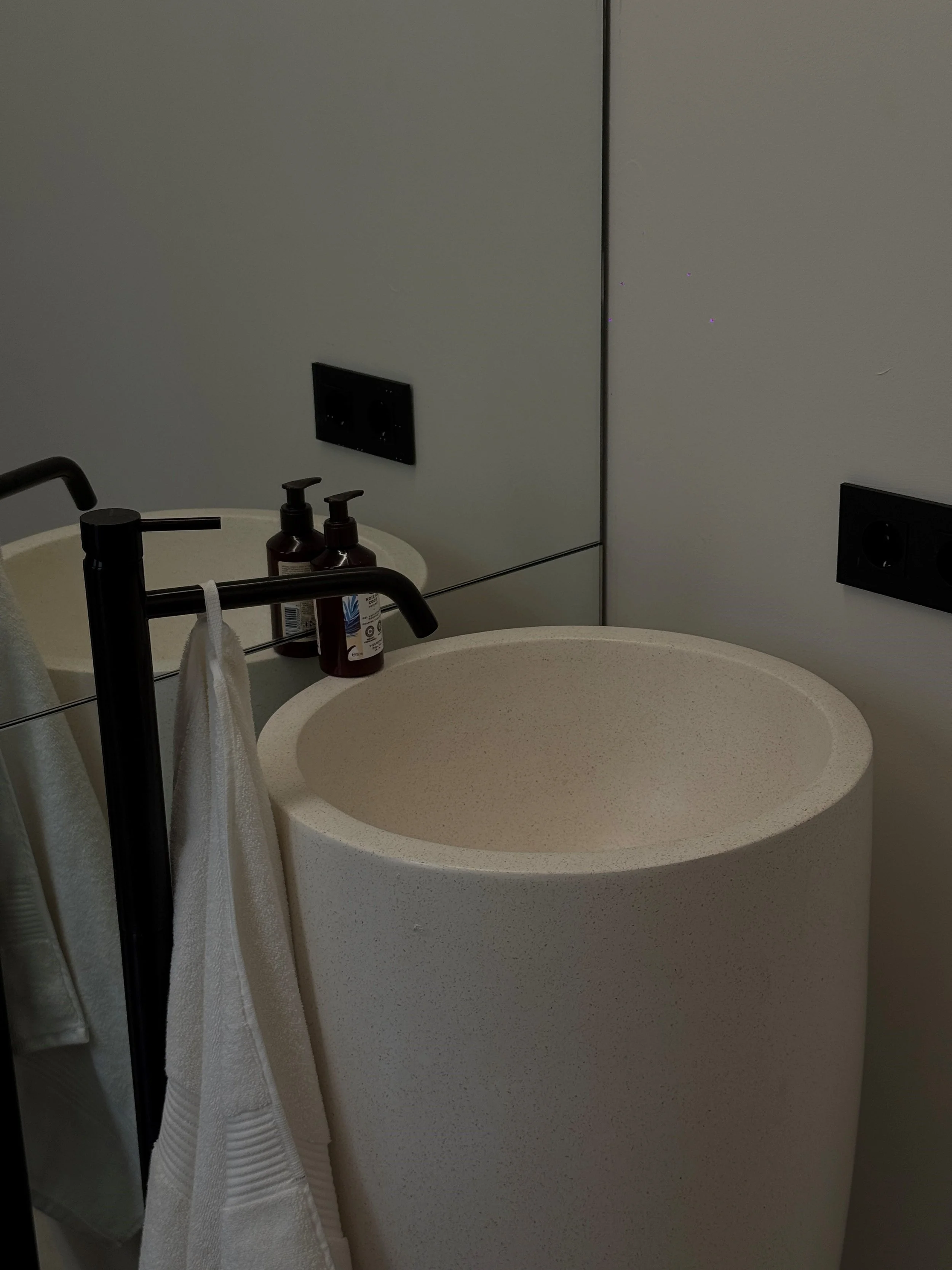 A minimalist beige round sink with black faucet, white towels hanging on a black towel rack, and two brown soap bottles on a white circular table, with black electrical outlets on a gray wall behind.