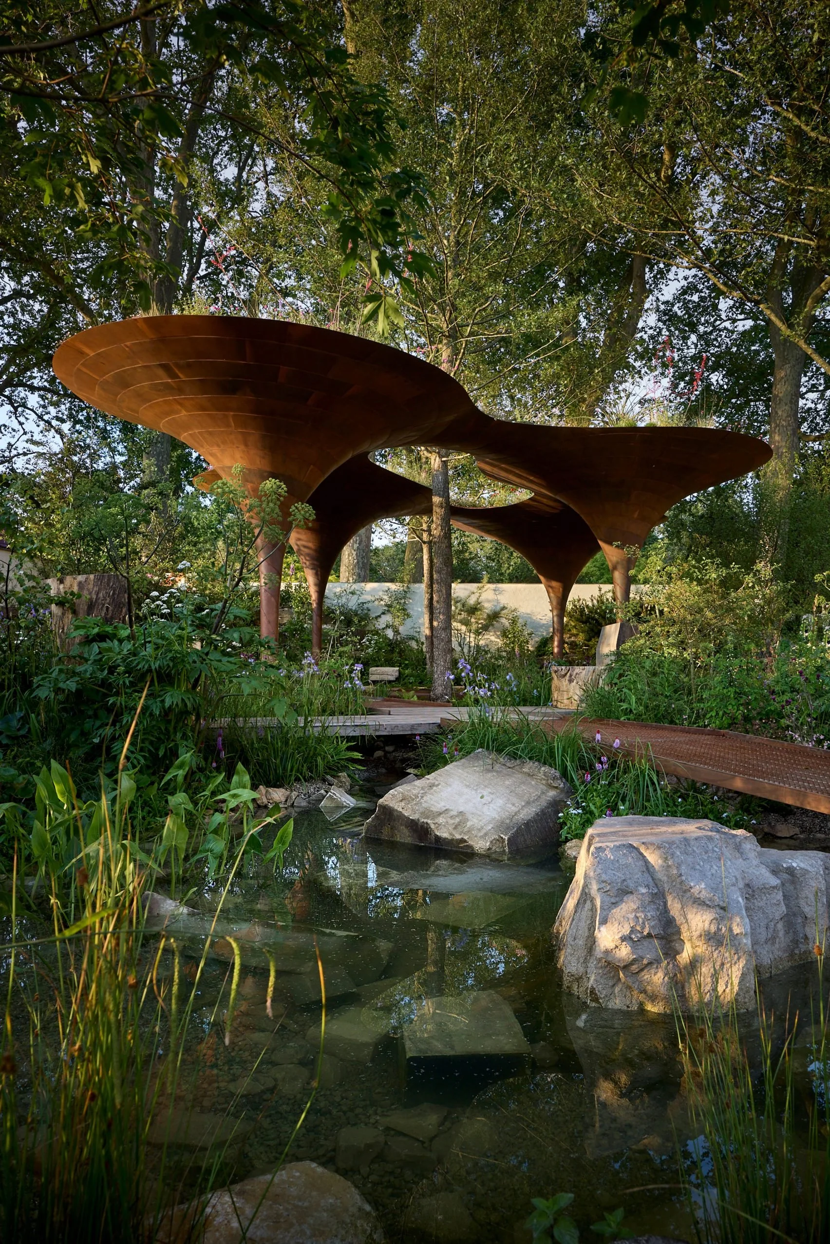 A garden with a pond, rocks, and lush green plants, featuring a unique wooden canopy structure with multiple curved, mushroom-like shapes.