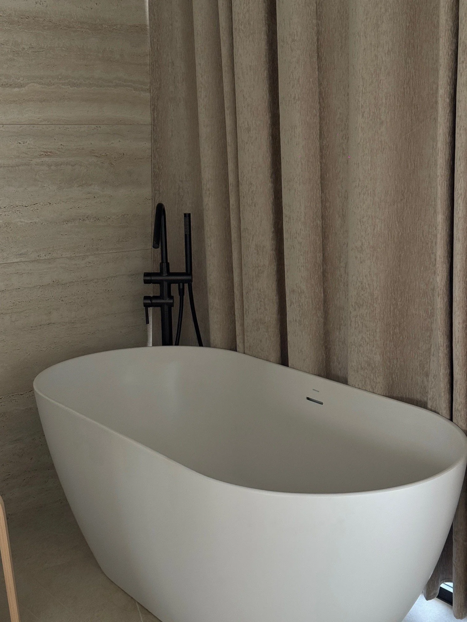 Modern white freestanding bathtub next to travertine walls and curtains, with black faucet and fixtures