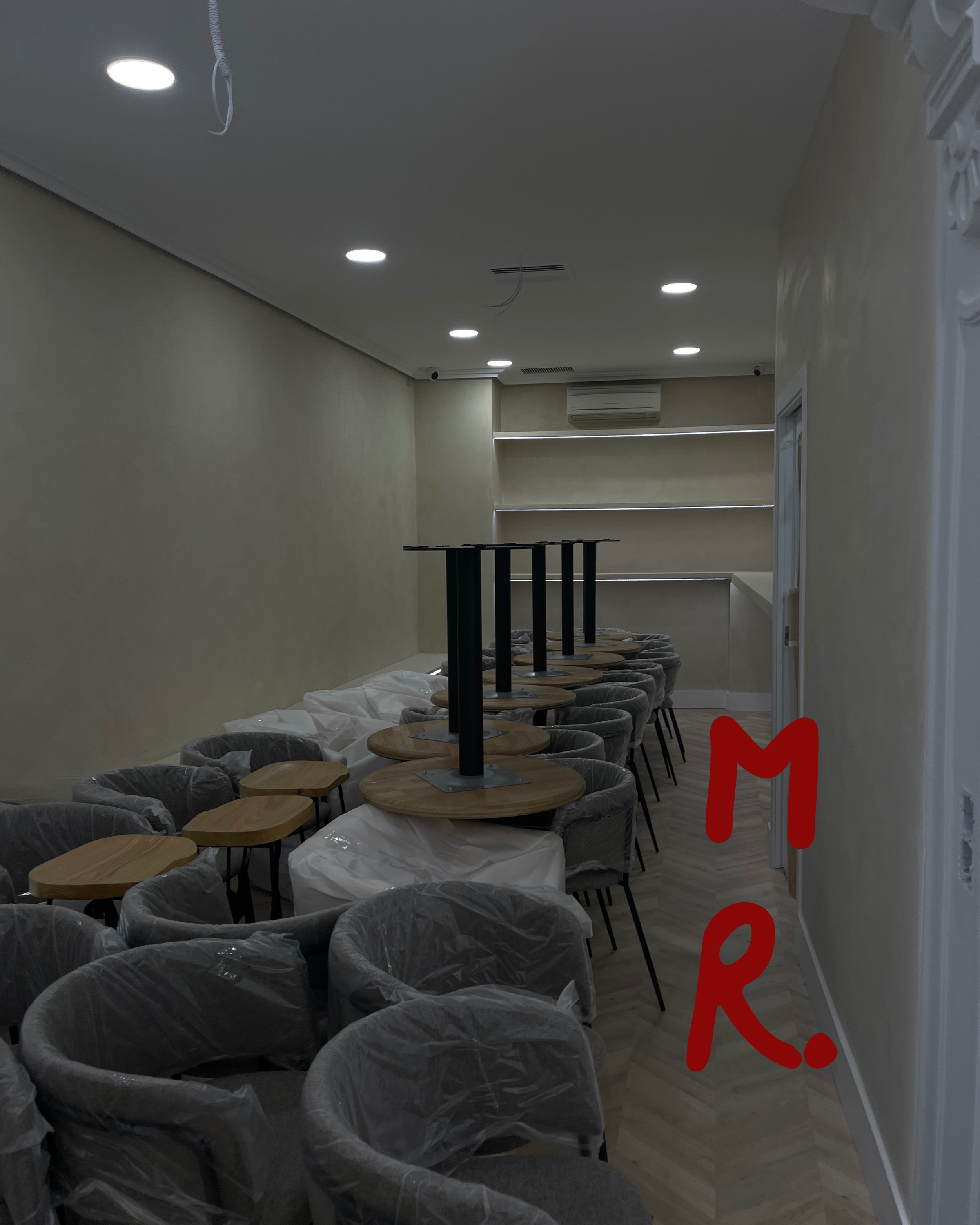 Empty restaurant or cafe interior with chairs covered in plastic and tables in the process of being assembled, ceiling lights, an air conditioning unit, and shelves on the back wall.