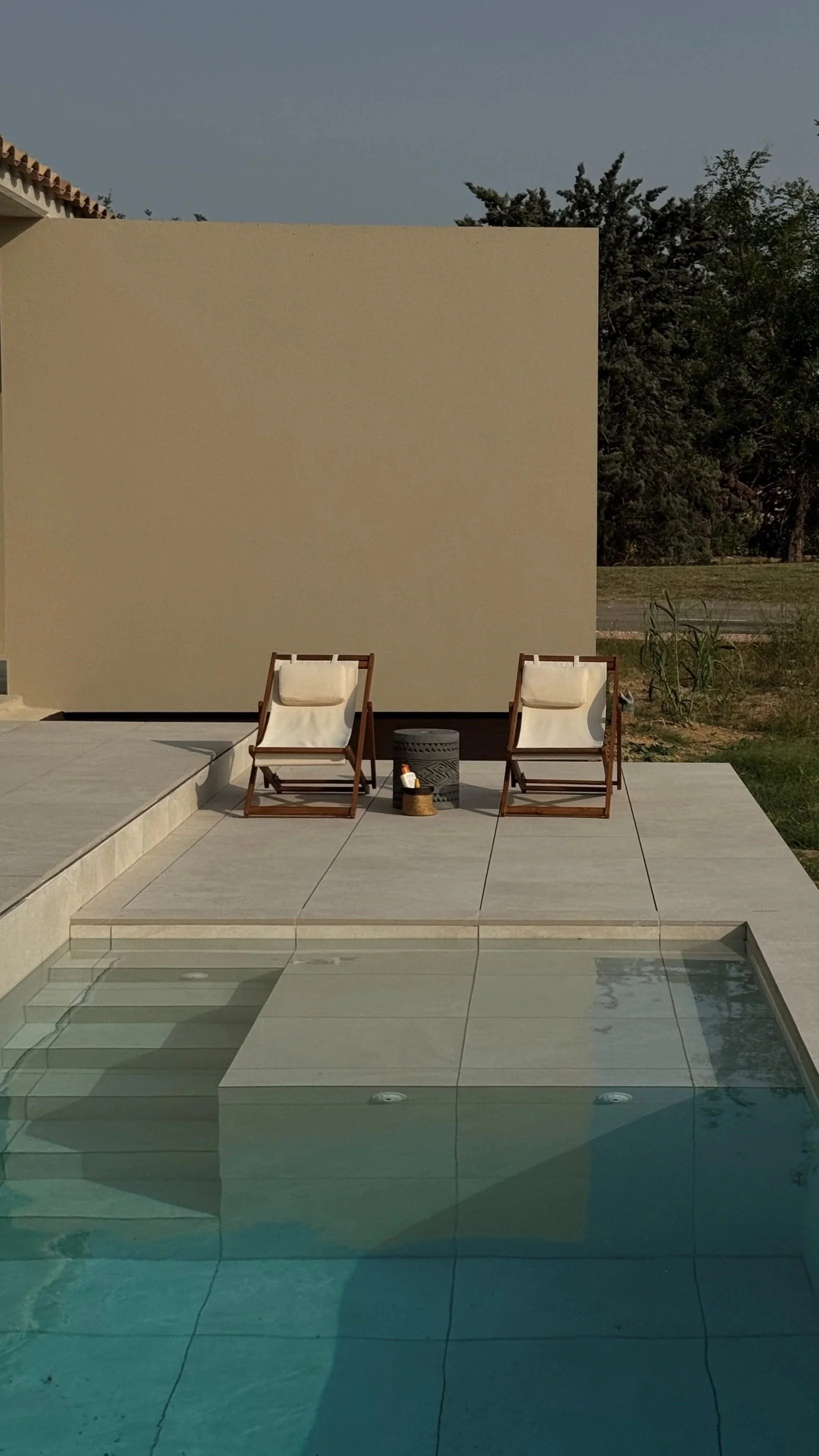 Two poolside lounge chairs with beige cushions and a small black cylindrical side table with a woven basket next to it, set on a tiled patio next to a swimming pool, with a beige privacy wall and greenery in the background.