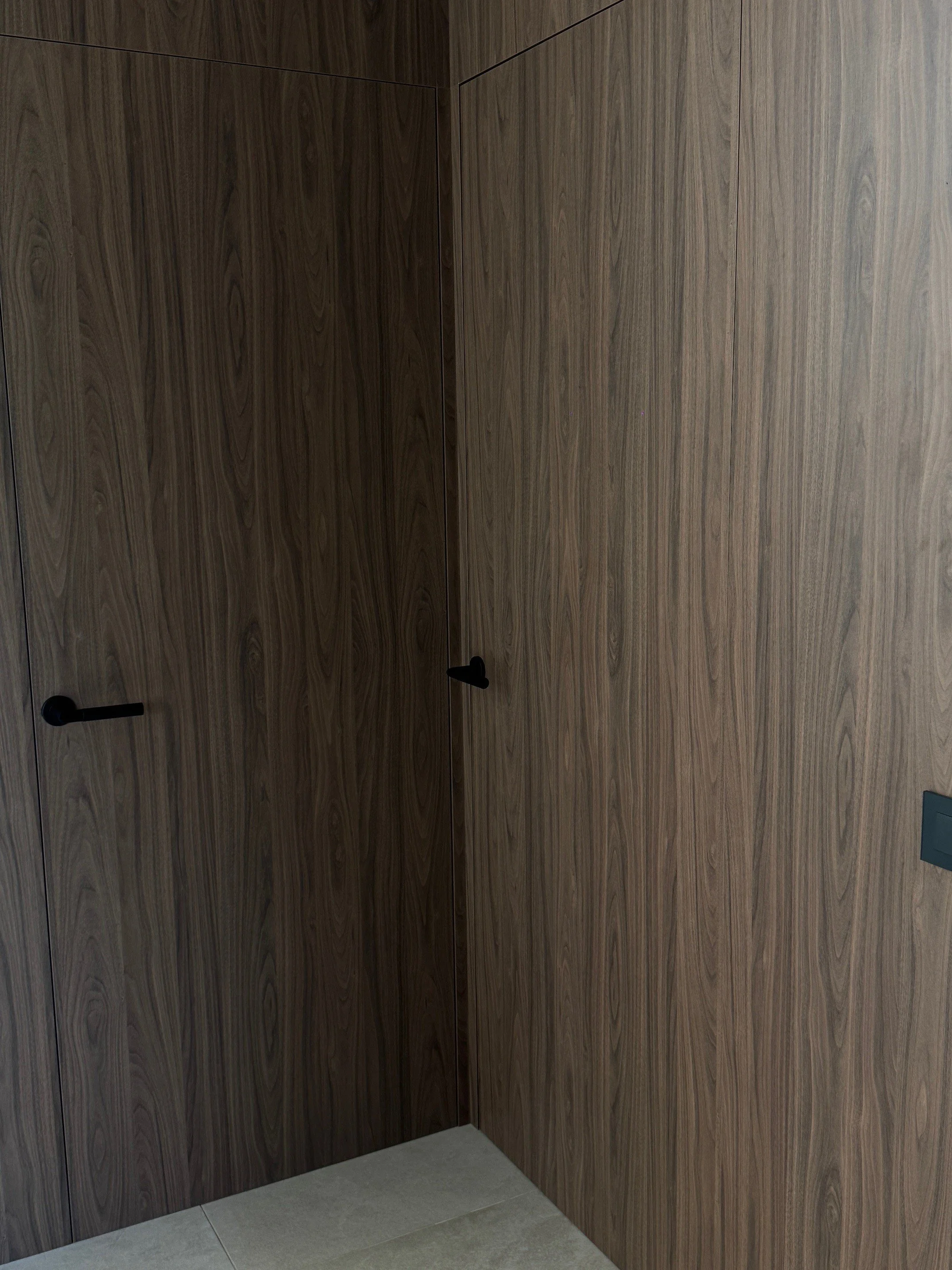 Wood-paneled walls with two black door handles on the left and a gray switch on the right wall, with beige tiled flooring.