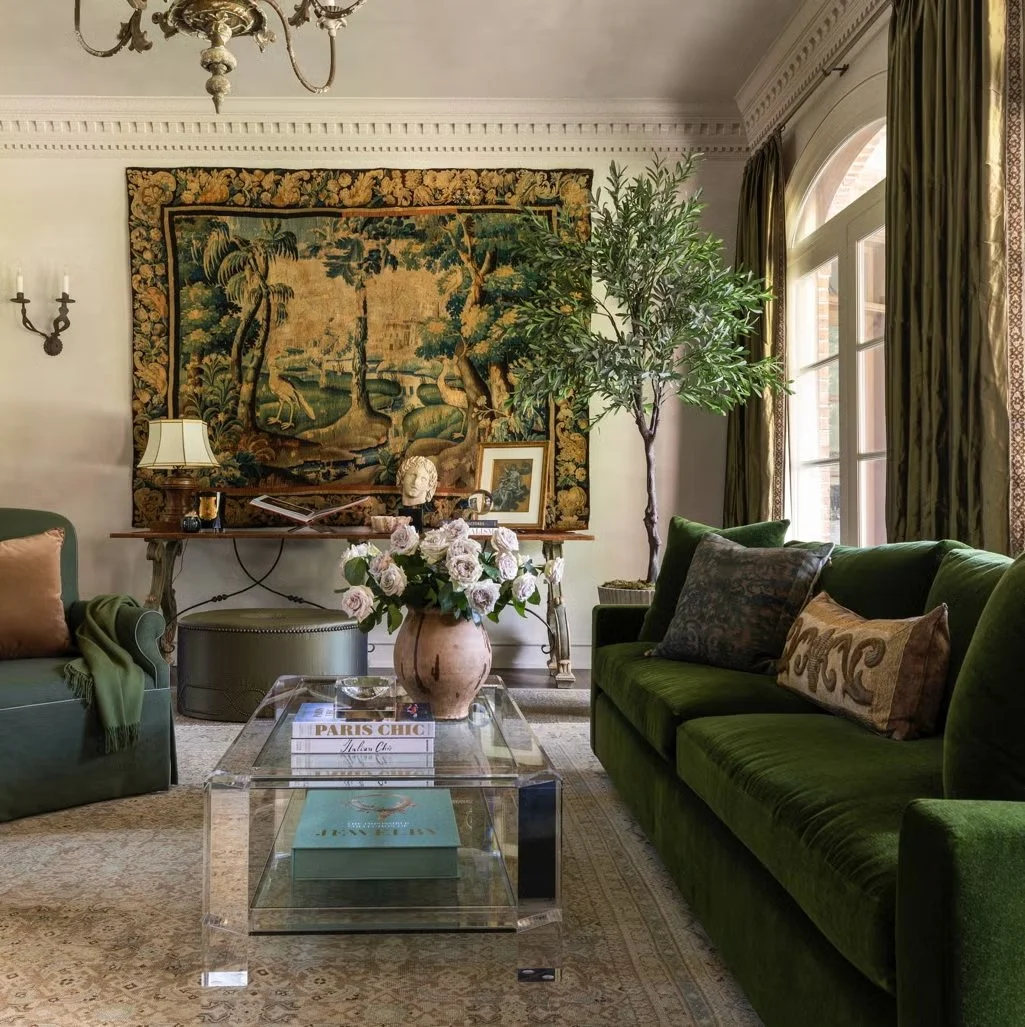 A living room with green velvet sofas, a glass coffee table with books and a flower vase, a large wall tapestry depicting a landscape, a potted tree, and a window with green curtains.