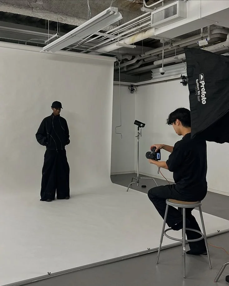 Photographer taking pictures of a model in a studio with a plain white backdrop, professional lighting equipment, and ceiling pipes.