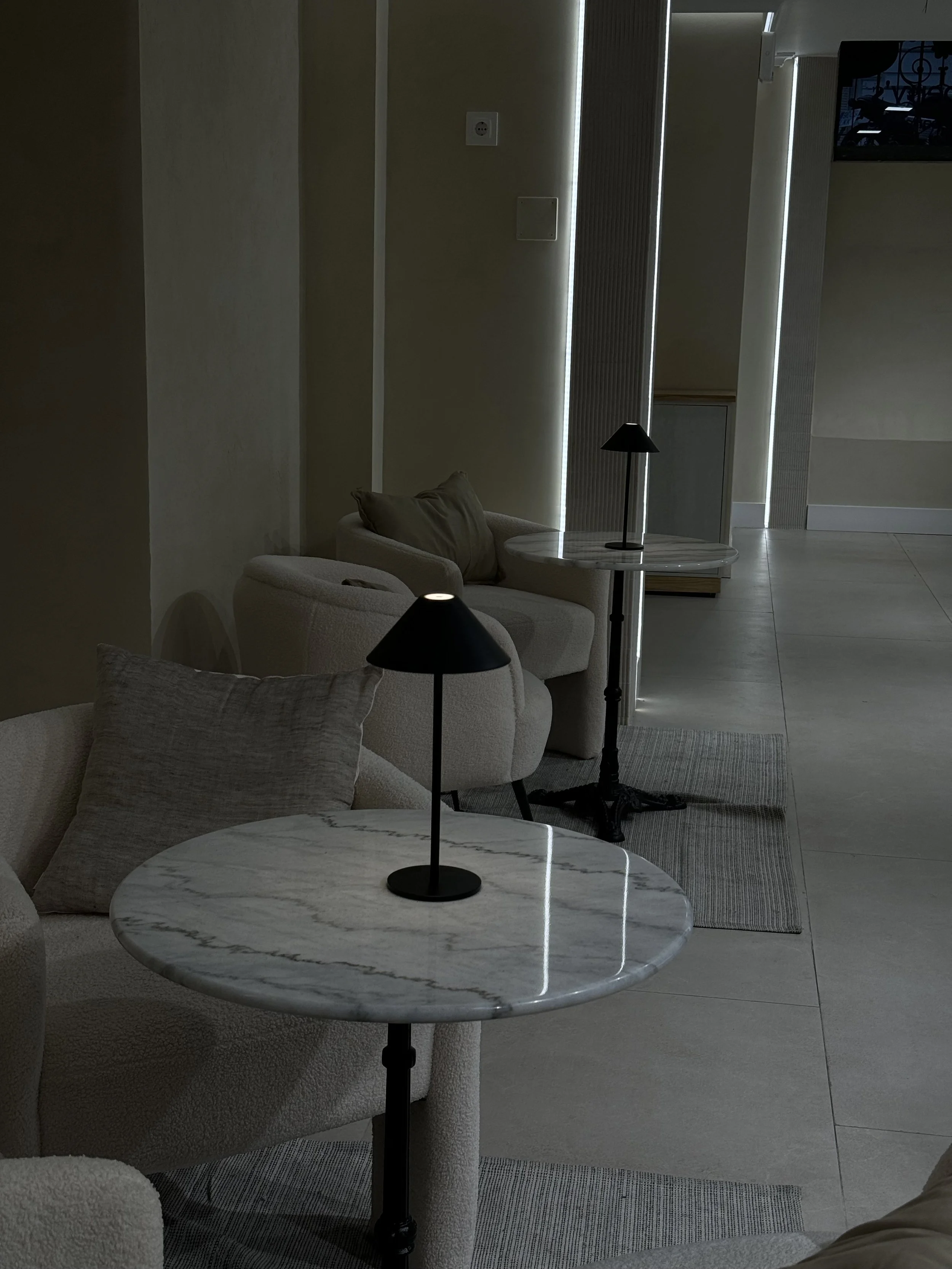 A modern, minimalist living room with beige sofas, marble-topped side tables with black lamps, a beige wall, and a doorway with vertical lighting strips.