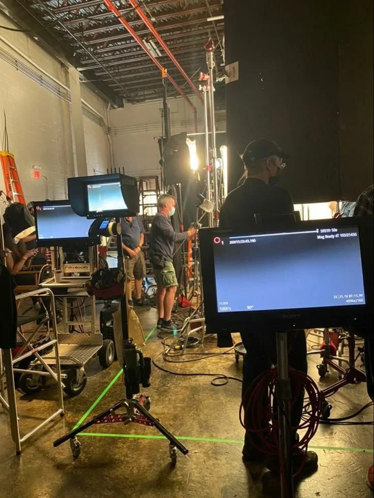 A behind-the-scenes look at a film or television production set with crew members, cameras, monitors, lighting equipment, and people working in a studio.