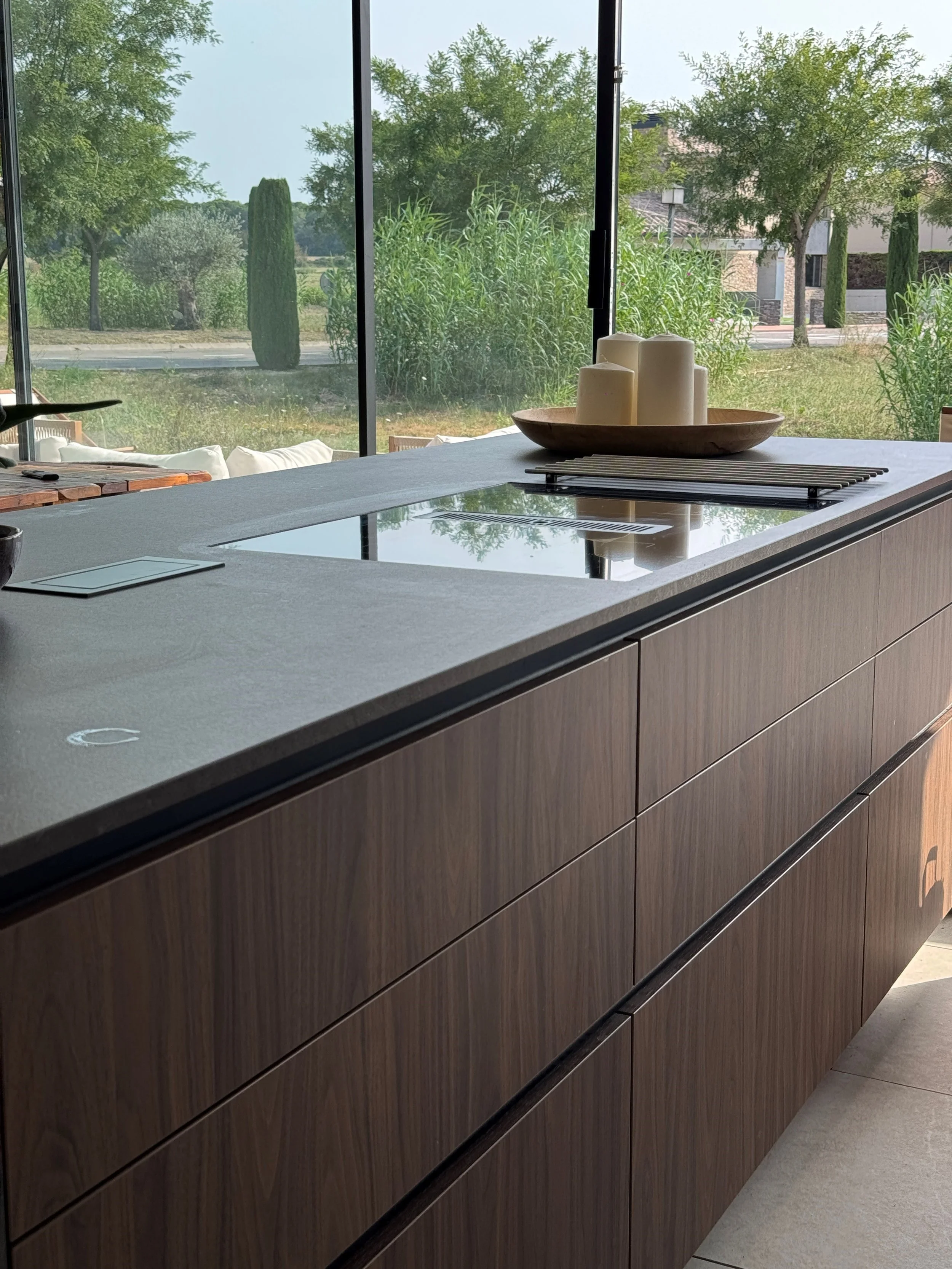 Modern kitchen counter with candles on a wooden tray, situated near large windows overlooking a green outdoor landscape.