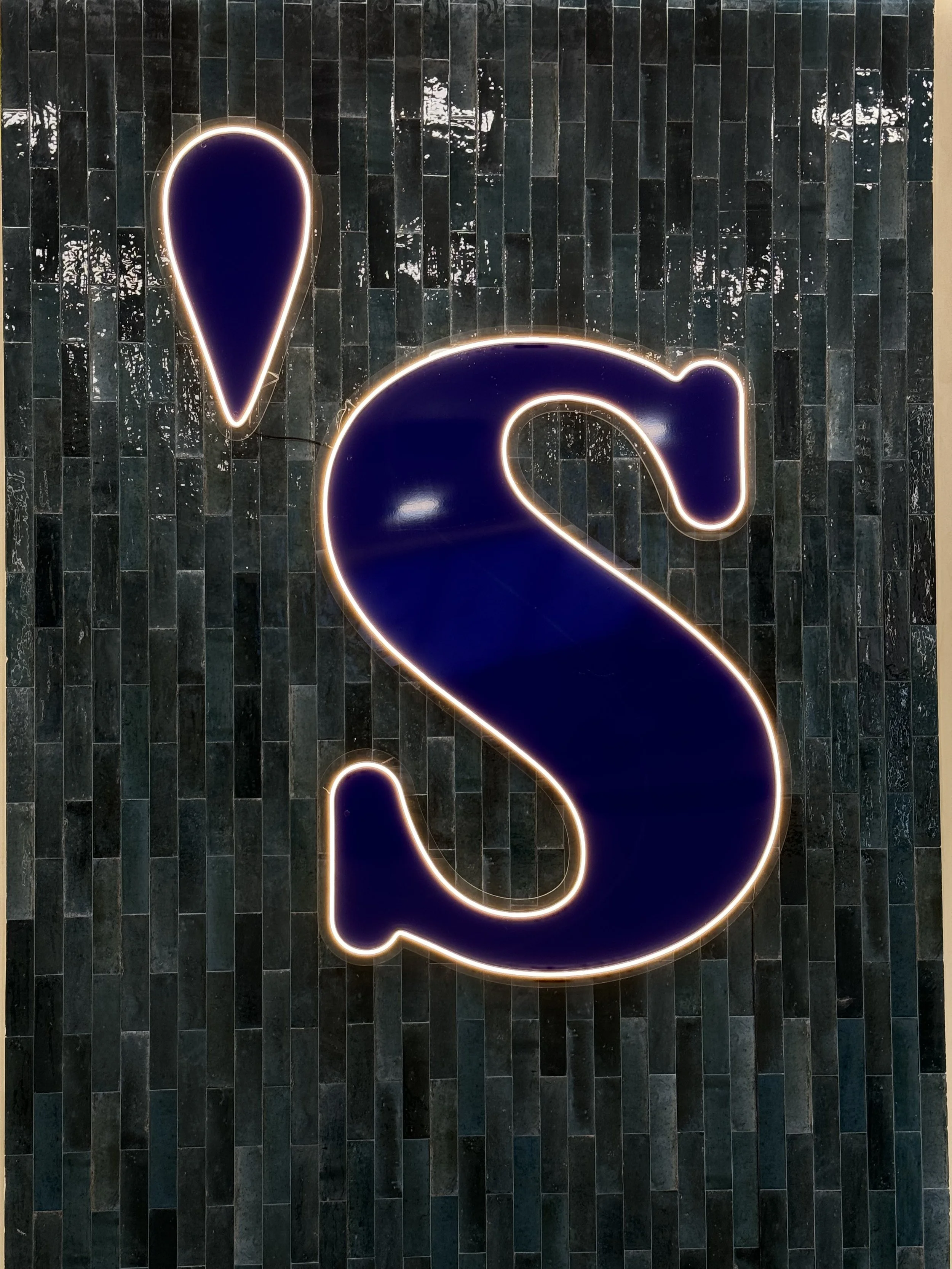 Illuminated neon sign of a large, dark blue letter 'S' with a smaller droplet-shaped icon above it, mounted on a black tiled wall.