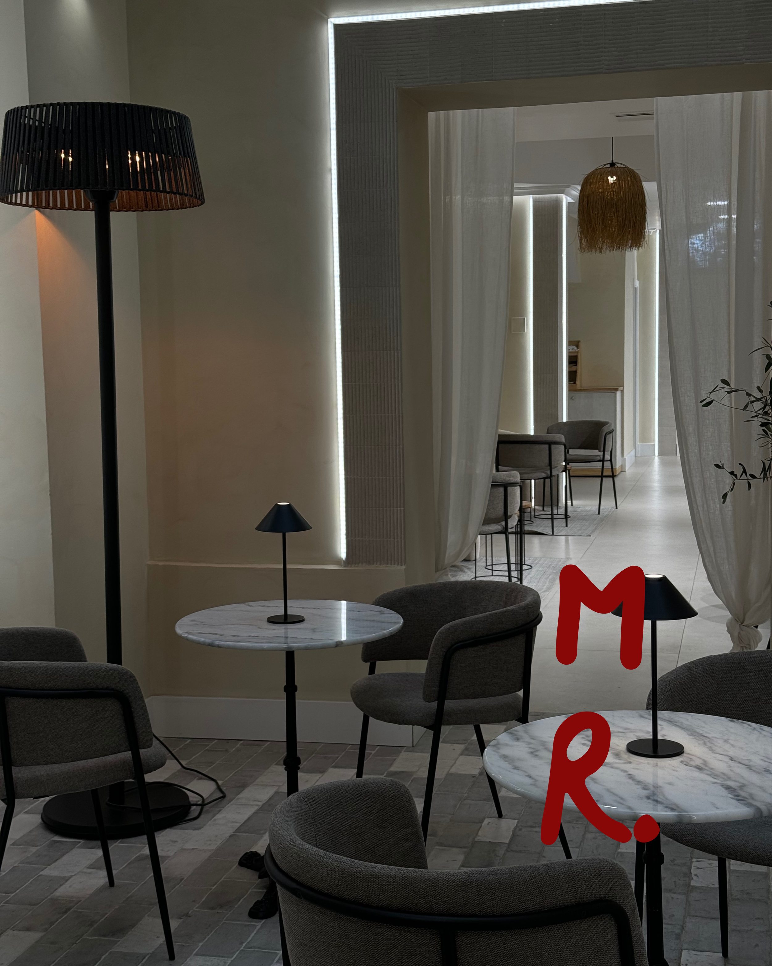 Interior of a modern cafe or lounge with round marble tables, gray upholstered chairs, small black table lamps, and ambient lighting, with curtains and additional seating visible in the background.