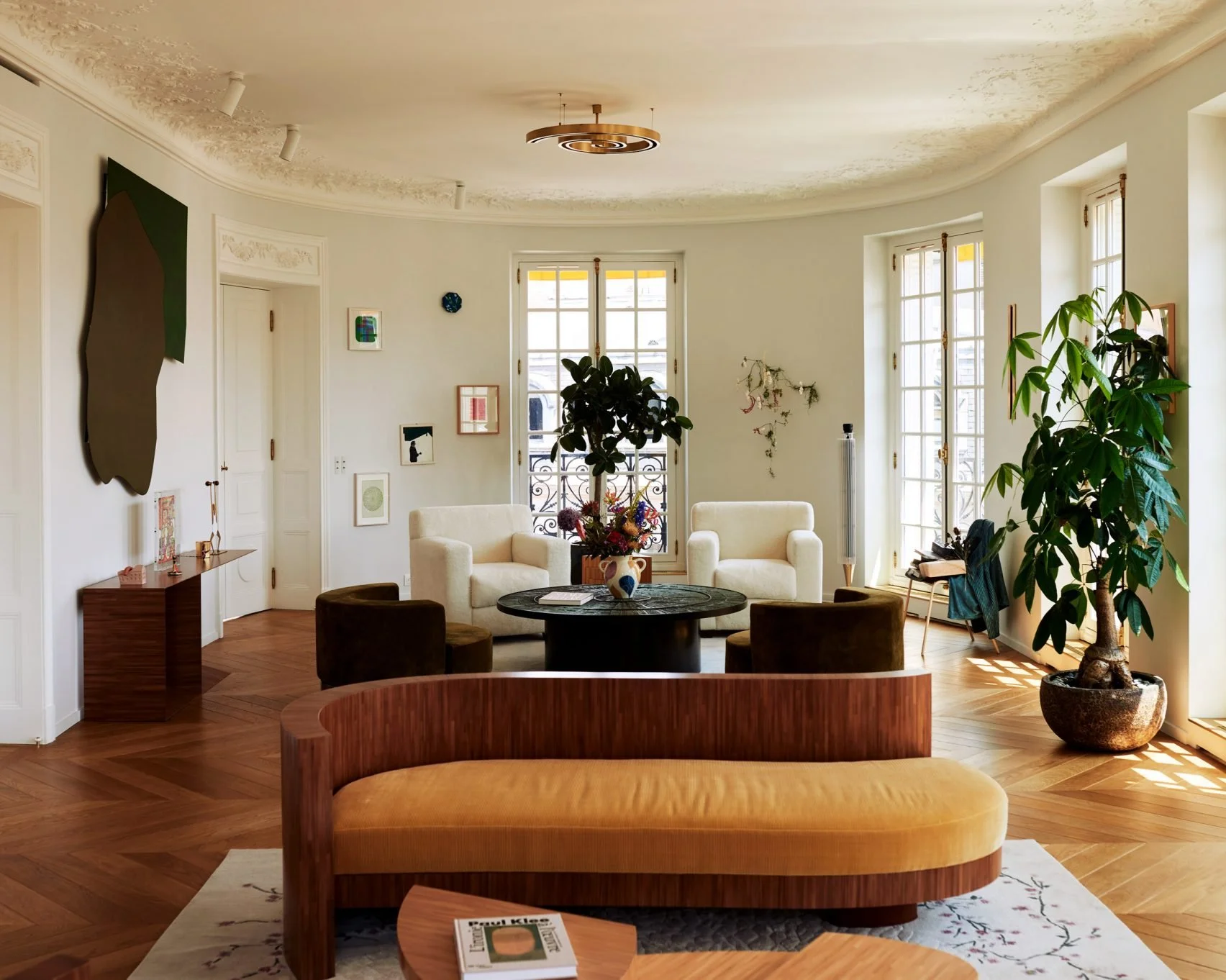 Living room with white walls, hardwood floors, large windows, and various seating including white and brown chairs and a wooden bench. Decor includes artwork, potted plants, a round table with a vase of flowers, and sunlight streaming in.