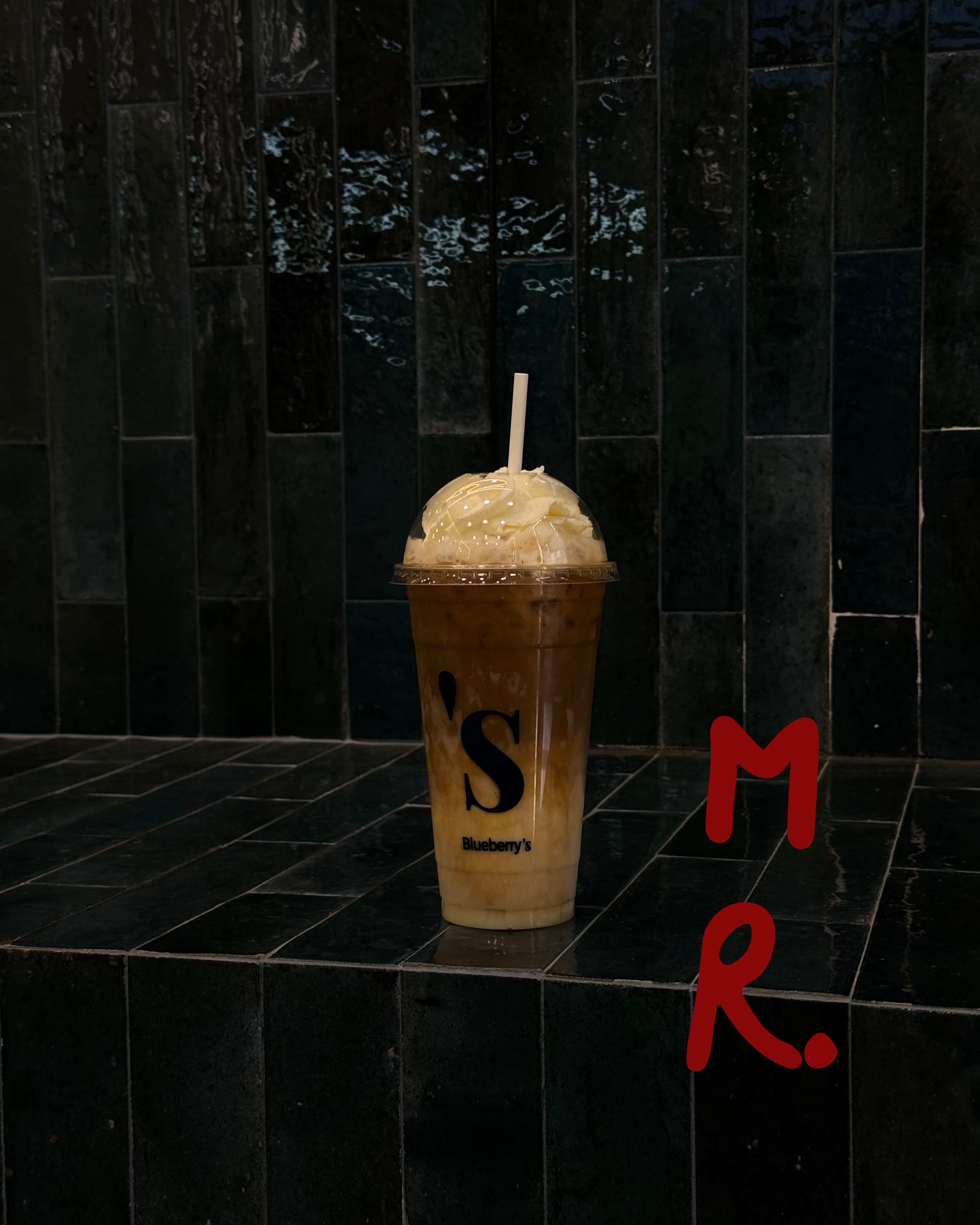 A large iced coffee drink with whipped cream on top in a clear plastic cup with a straw and the text 'S Blueberry's' on the side, placed on a black-tiled surface against a dark tiled wall, with red handwritten letters 'M R.' on the right side of the 