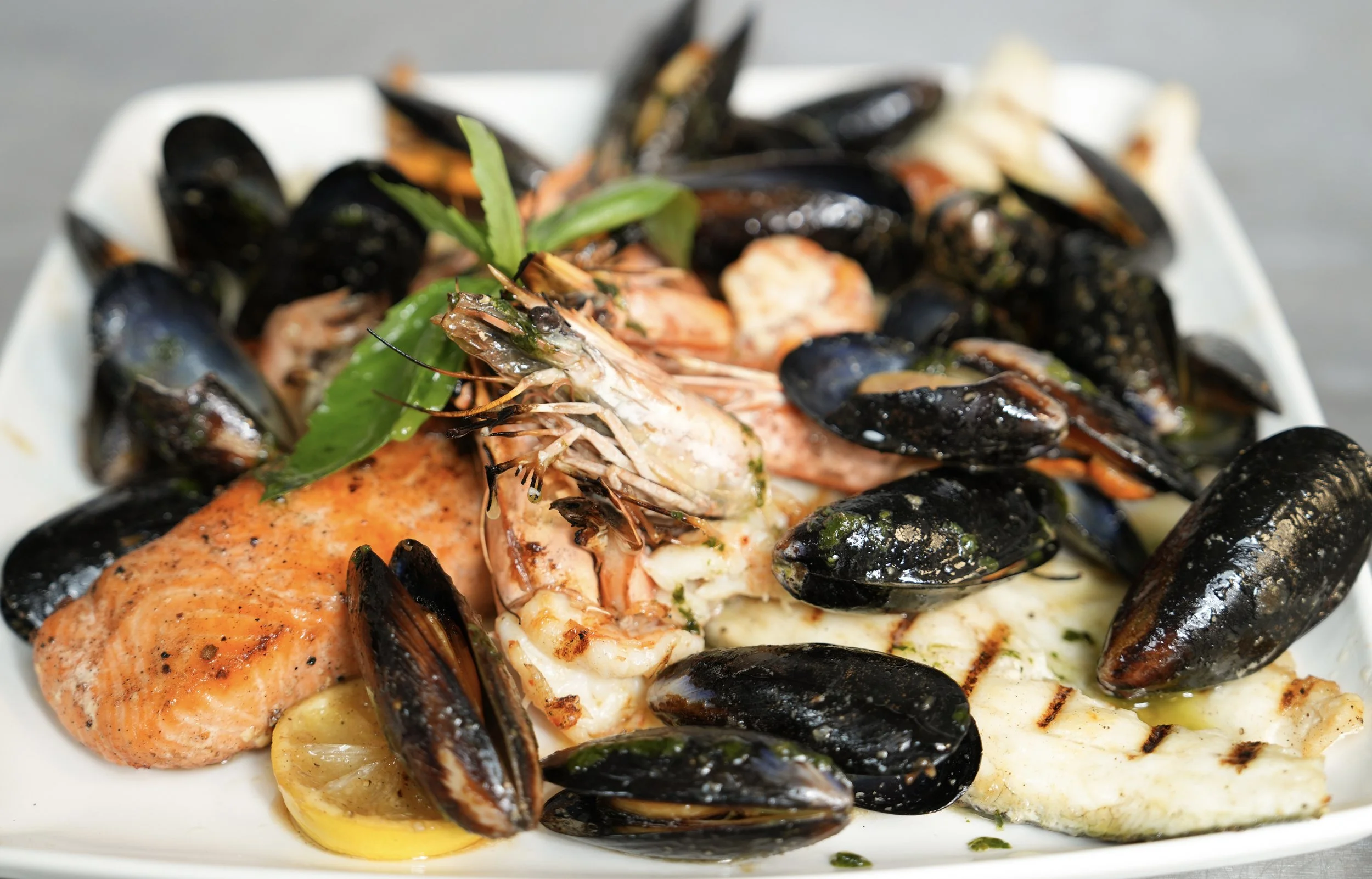 Seafood platter with grilled salmon, shrimp, and mussels garnished with herbs and lemon slices on a white plate.