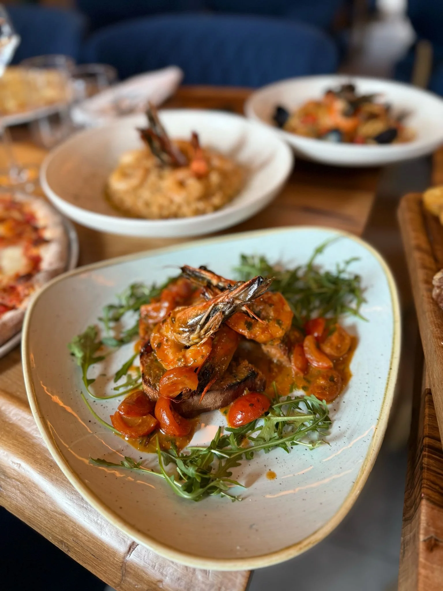 A gourmet meal featuring grilled shrimp on a bed of arugula and sliced cherry tomatoes on a white plate. In the background, a bowl of risotto and another dish are visible.