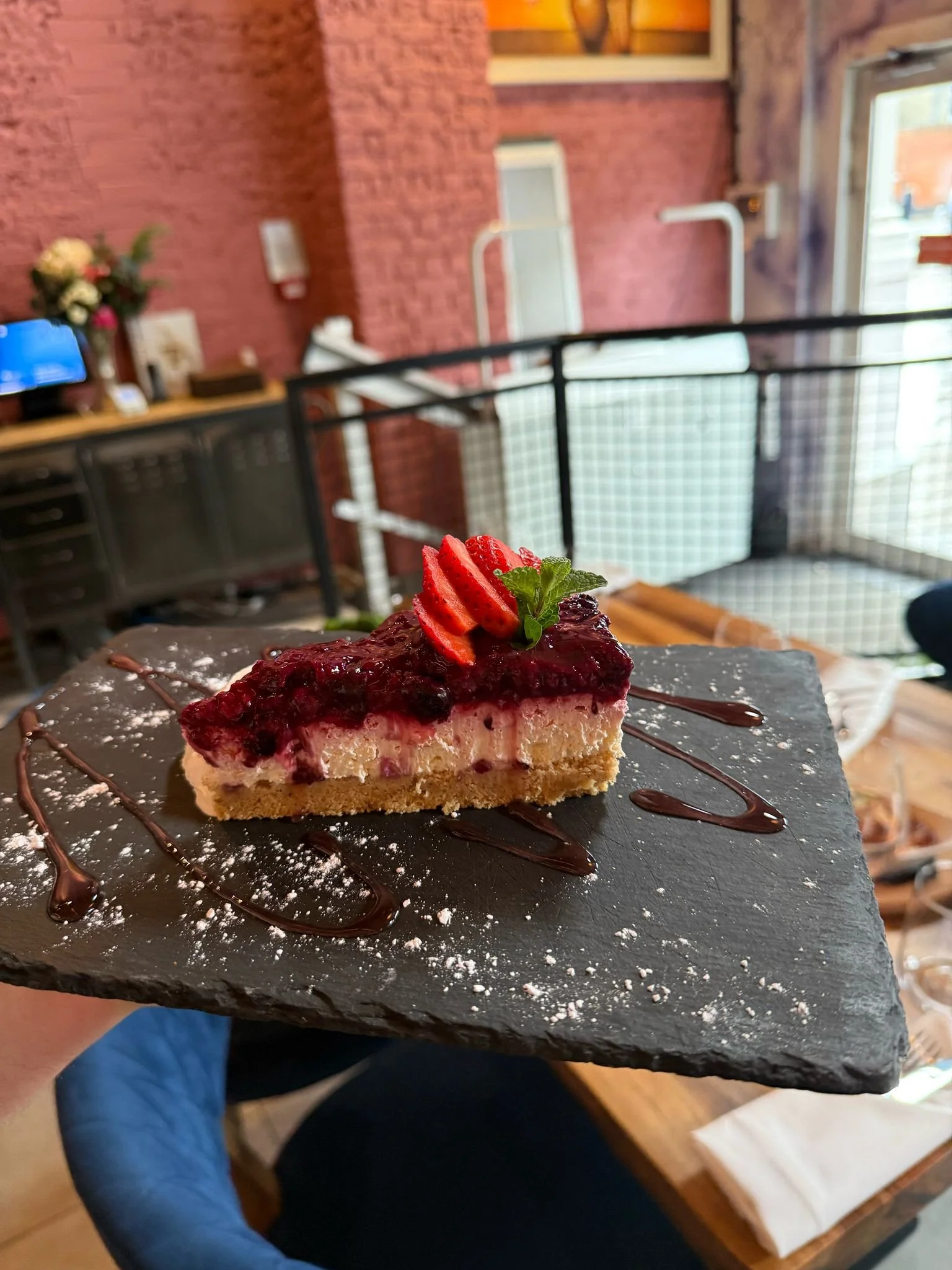 Berry cheesecake with strawberry slices on a slate plate, garnished with mint and chocolate drizzle.