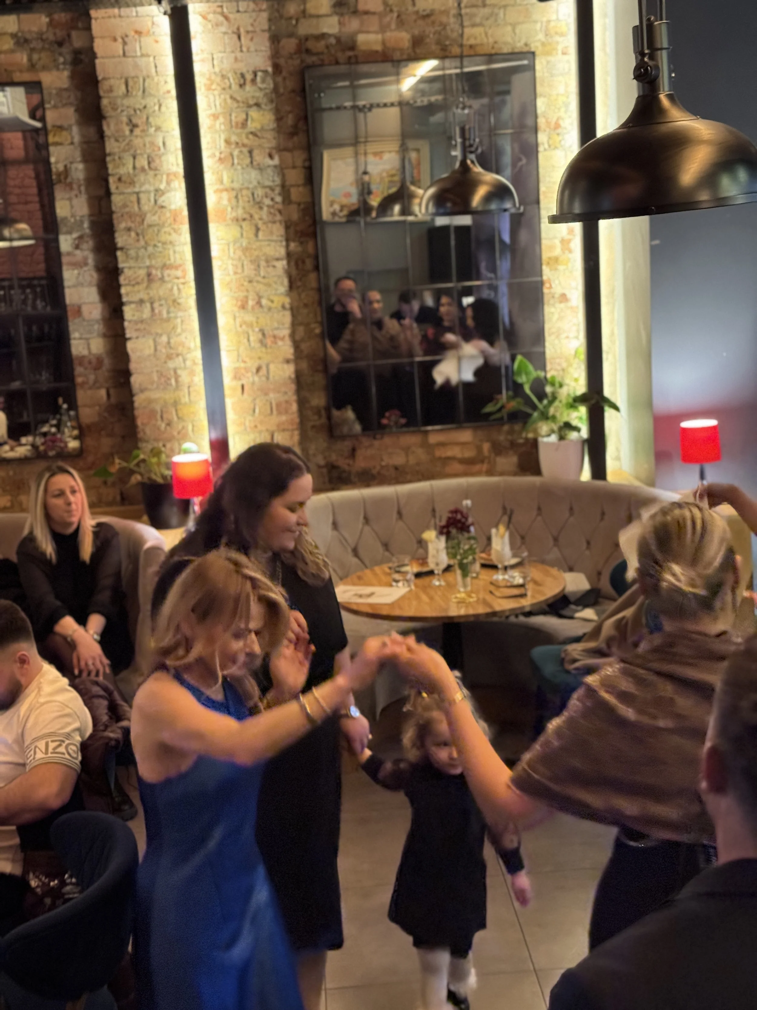 People dancing in a cozy restaurant with exposed brick walls and soft lighting. A small child joins the adults in a circle dance near a table. Reflections are visible in a wall mirror.