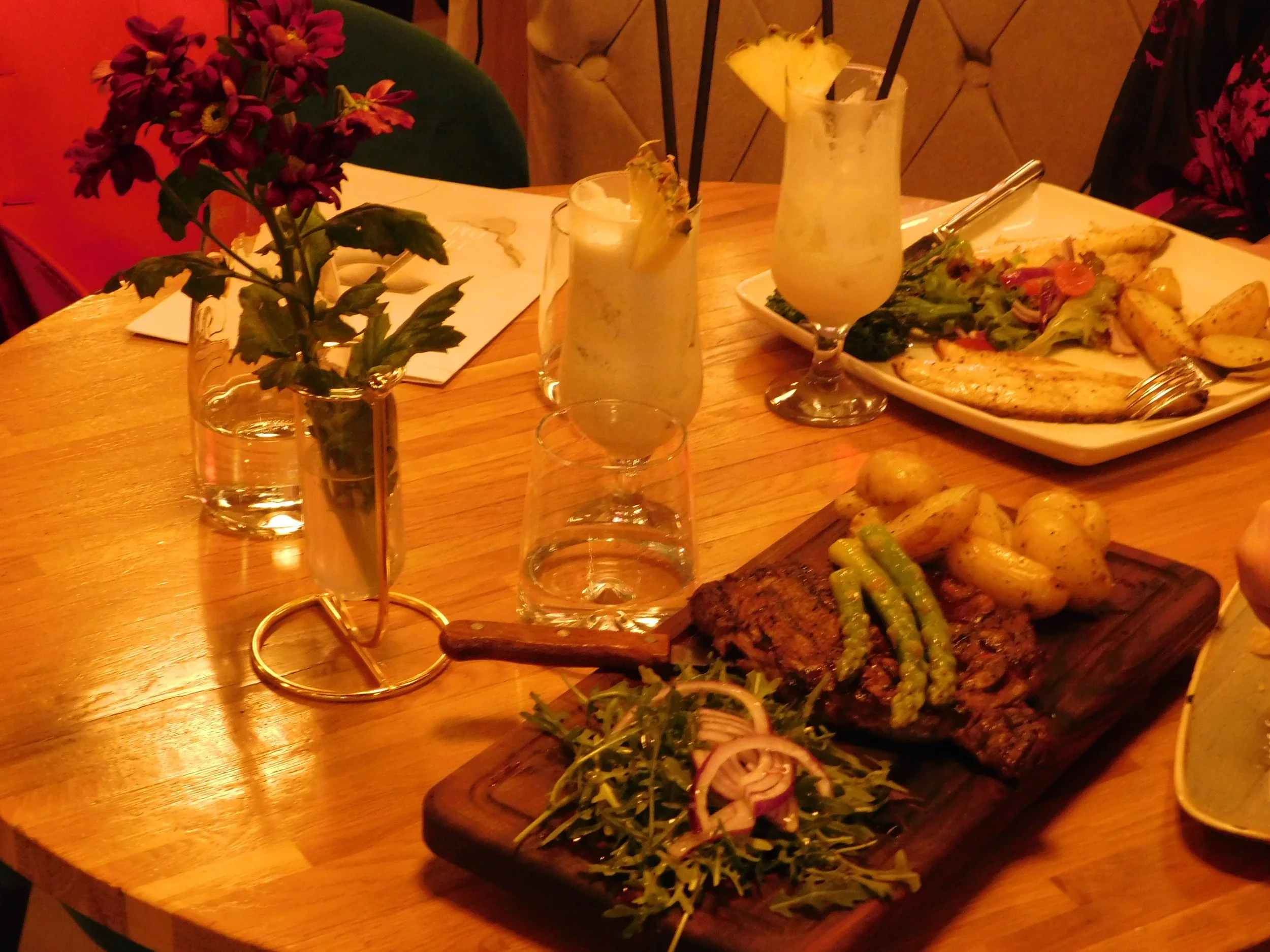 Plate with grilled steak, potatoes, and asparagus on a wooden board, next to a floral vase and a drink with a pineapple slice on a wooden table.