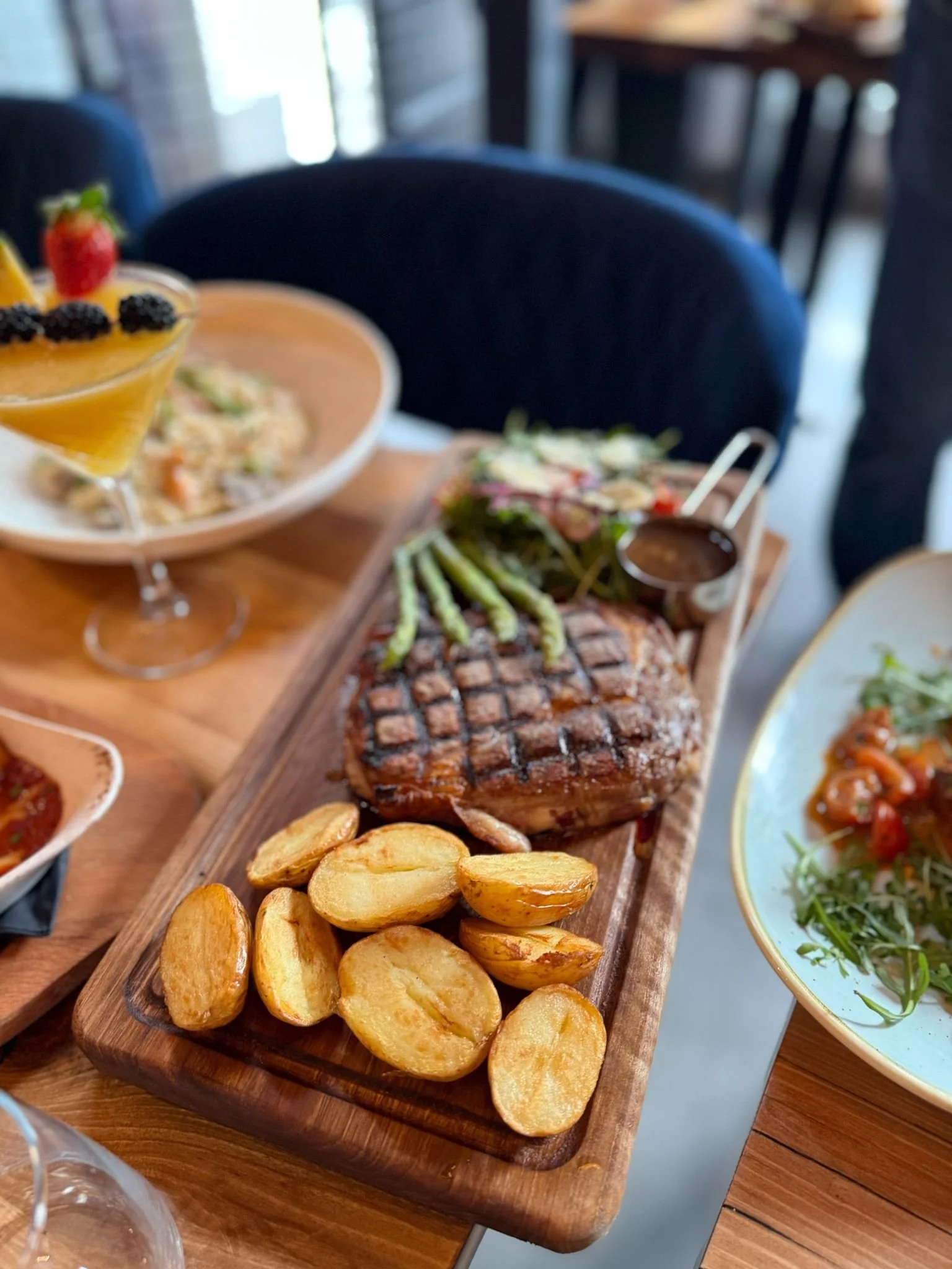 Restaurant table setting with grilled steak, roasted potatoes, asparagus, salad, and a cocktail with fruit garnish.