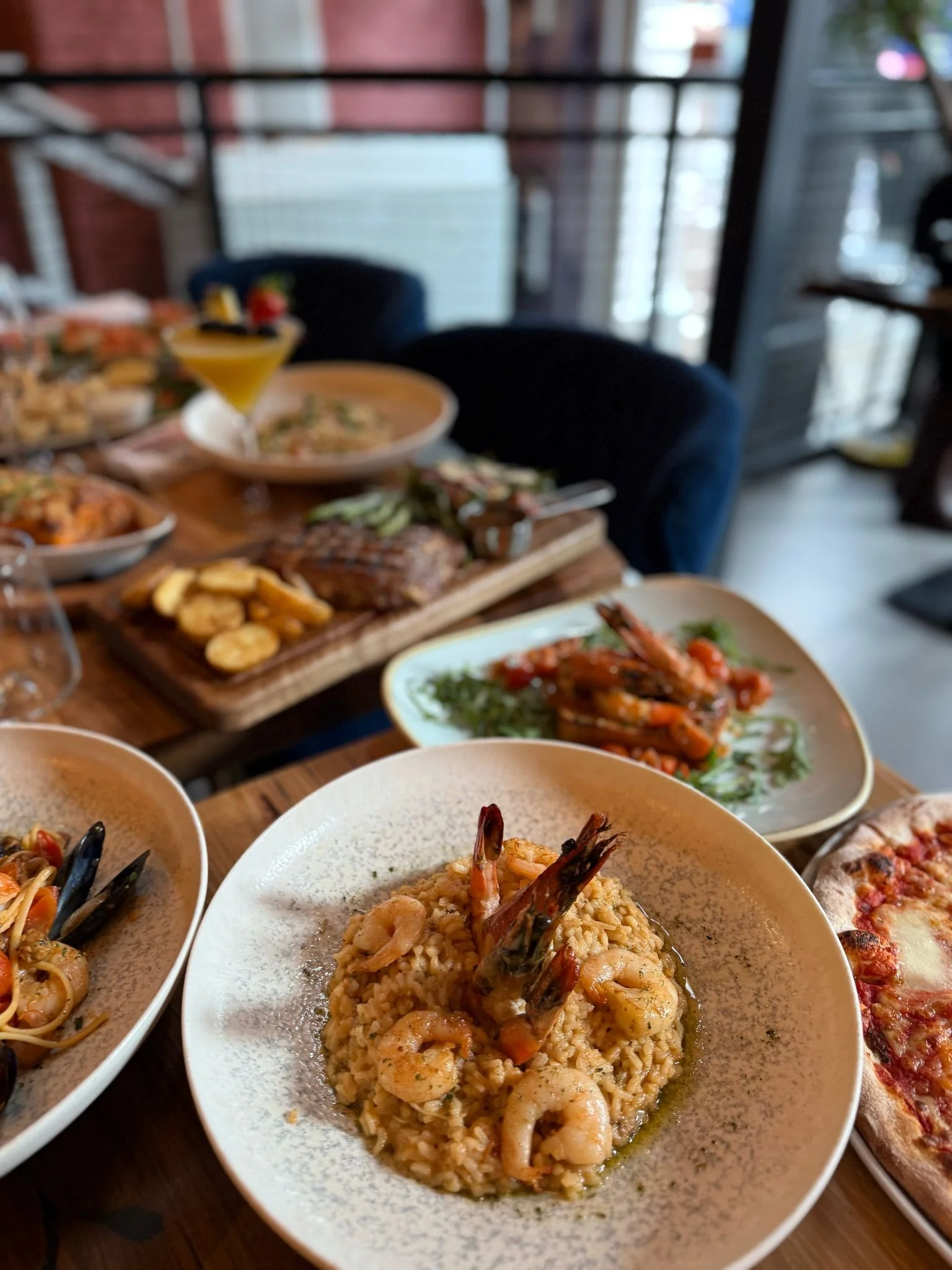 An assortment of dishes including shrimp risotto, seafood pasta with mussels, grilled shrimp, pizza, and a cocktail on a restaurant table.