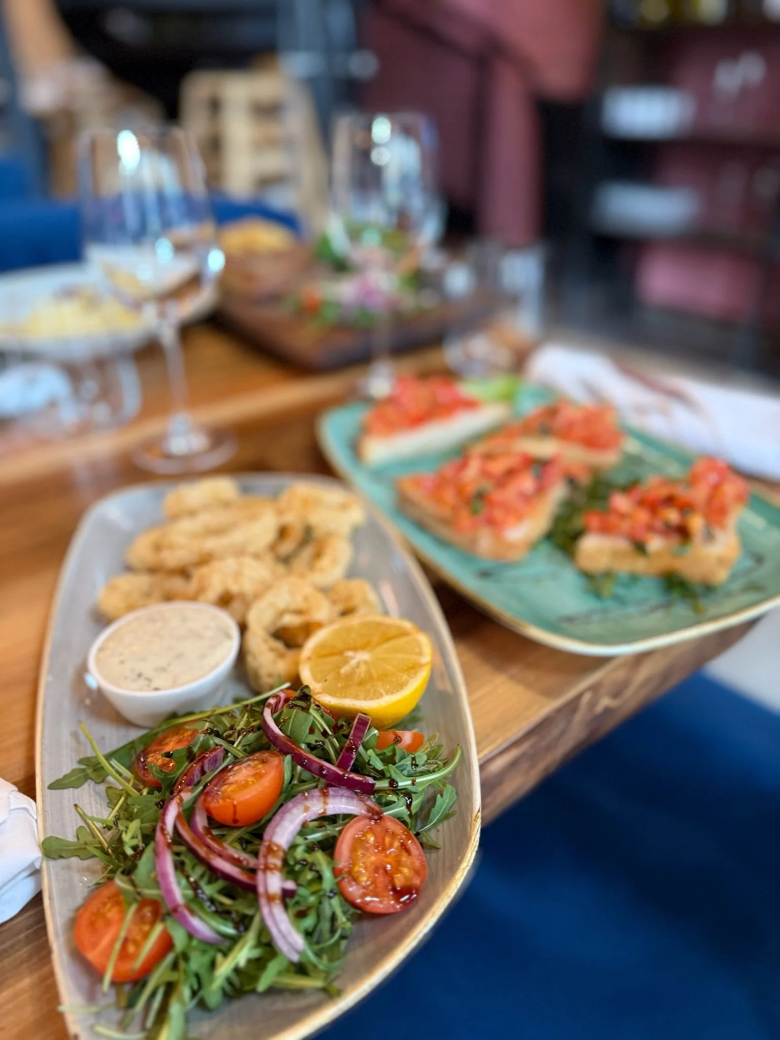 A plate of fried calamari with a lemon wedge, dipping sauce, and a fresh salad with arugula, cherry tomatoes, and red onion, served on a wooden table with wine glasses.