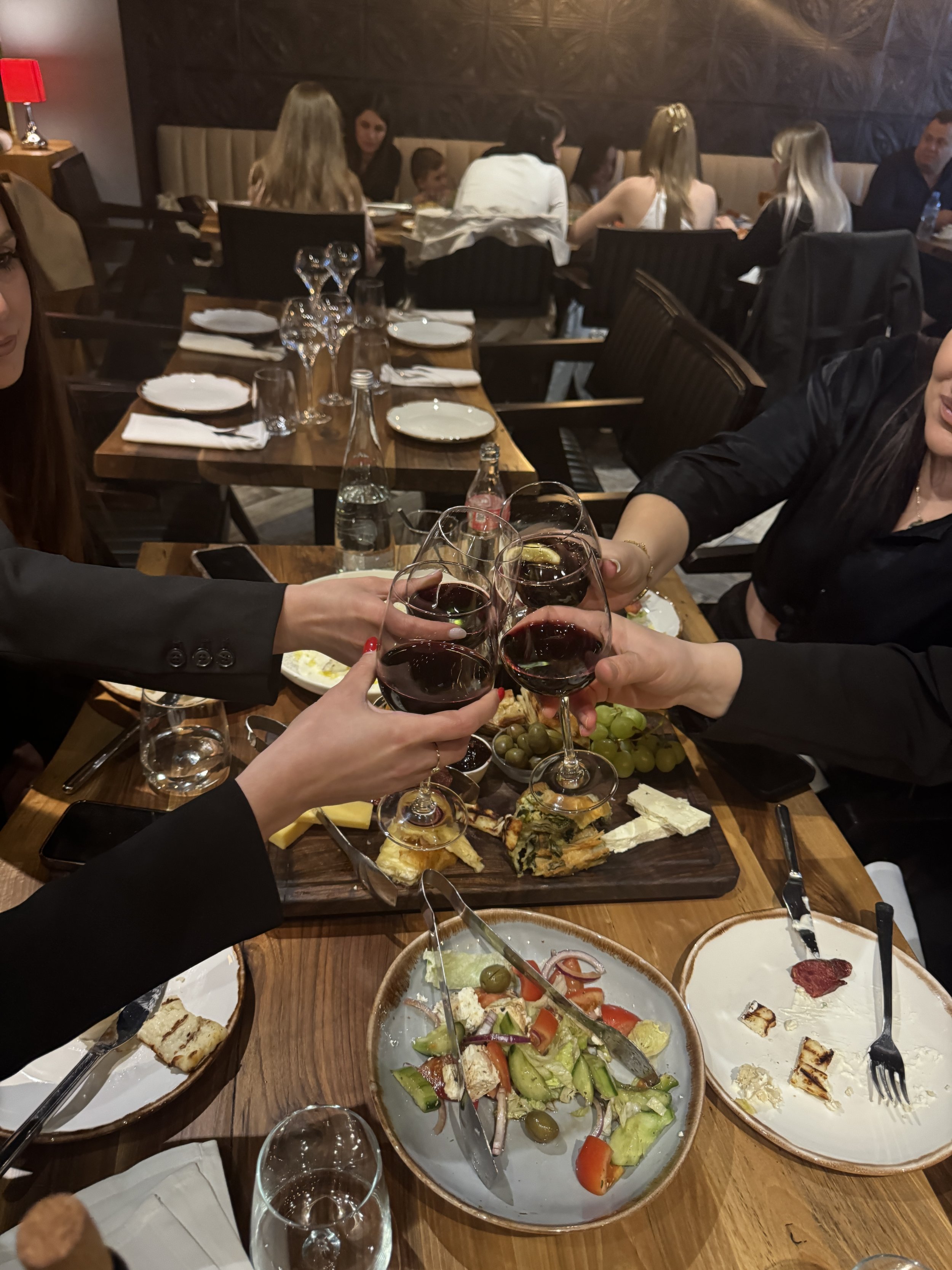 Group of people toasting with glasses of red wine over a table filled with assorted foods including salad, cheese, and bread.
