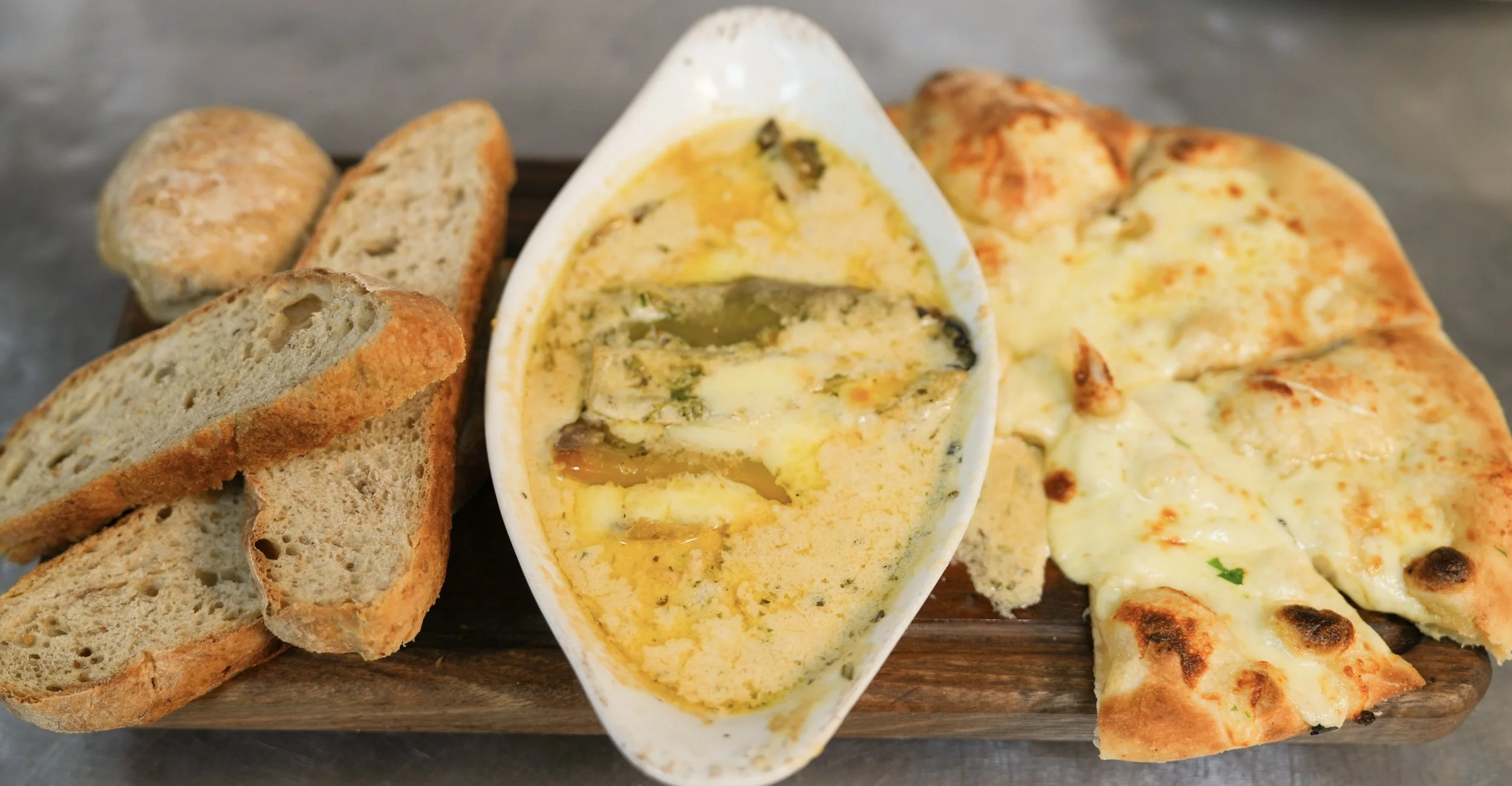 A dish of melted cheese fondue with slices of bread and cheese pizza on a wooden board.