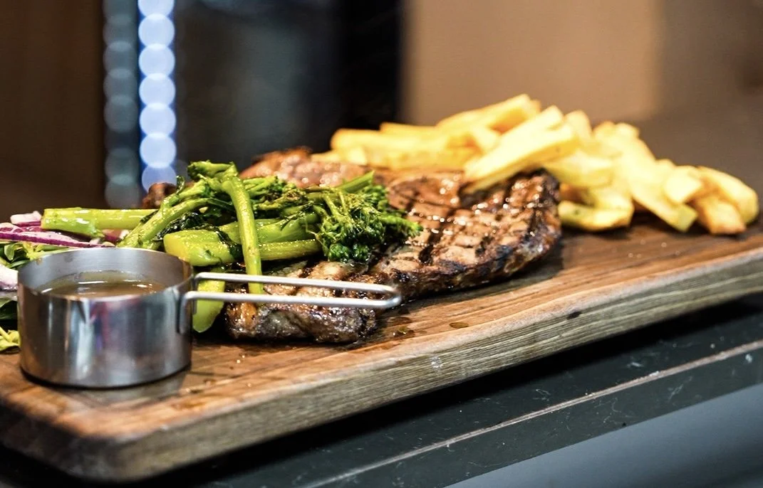 Grilled steak with broccolini, fries, and sauce on a wooden board.