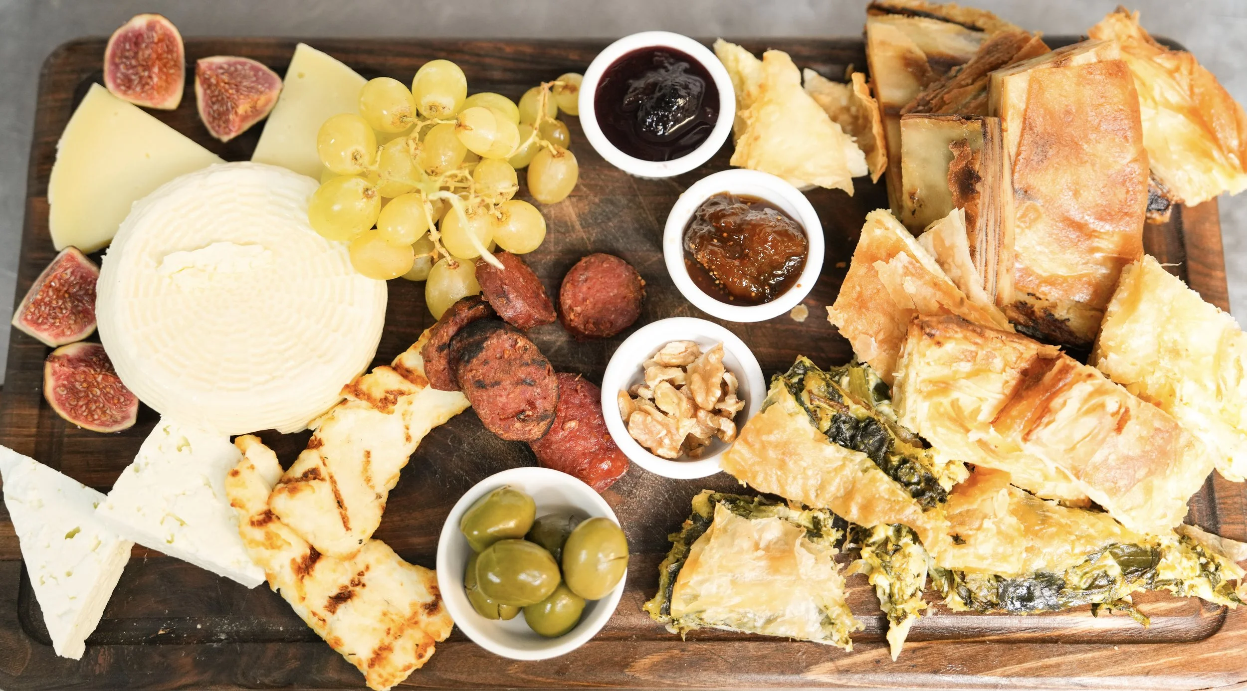 A charcuterie board with various cheeses, figs, grapes, sliced cured meats, nuts, olives, pastries, and small dishes of jam on a wooden surface.
