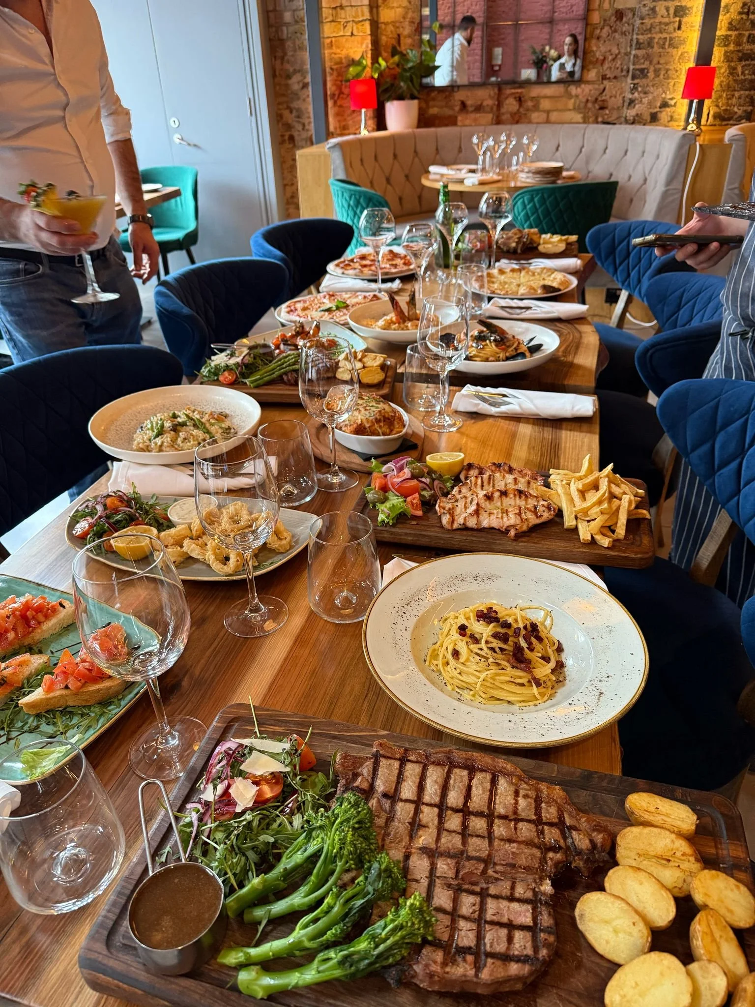 A dining table with various Italian dishes, including pasta, steak with greens and potatoes, bruschetta, grilled chicken with fries, and seafood. The table is set with wine glasses and surrounded by blue chairs. A person holding a drink stands nearby